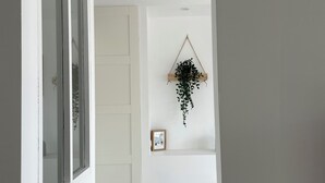 Une chambre avec un lit blanc, une fenêtre avec des stores, une porte et une plante fixée au mur.