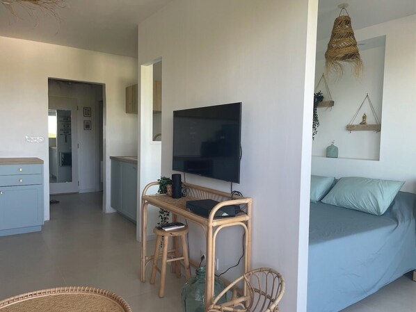 A modern, well-lit room with a large flat-screen TV mounted on the wall, a comfortable bed with light blue bedding, a small wooden desk with a chair, and a wicker chair.