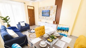 Una sala moderna con un sofá, sillones, una mesa de comedor preparada para cuatro personas y un televisor montado en la pared.