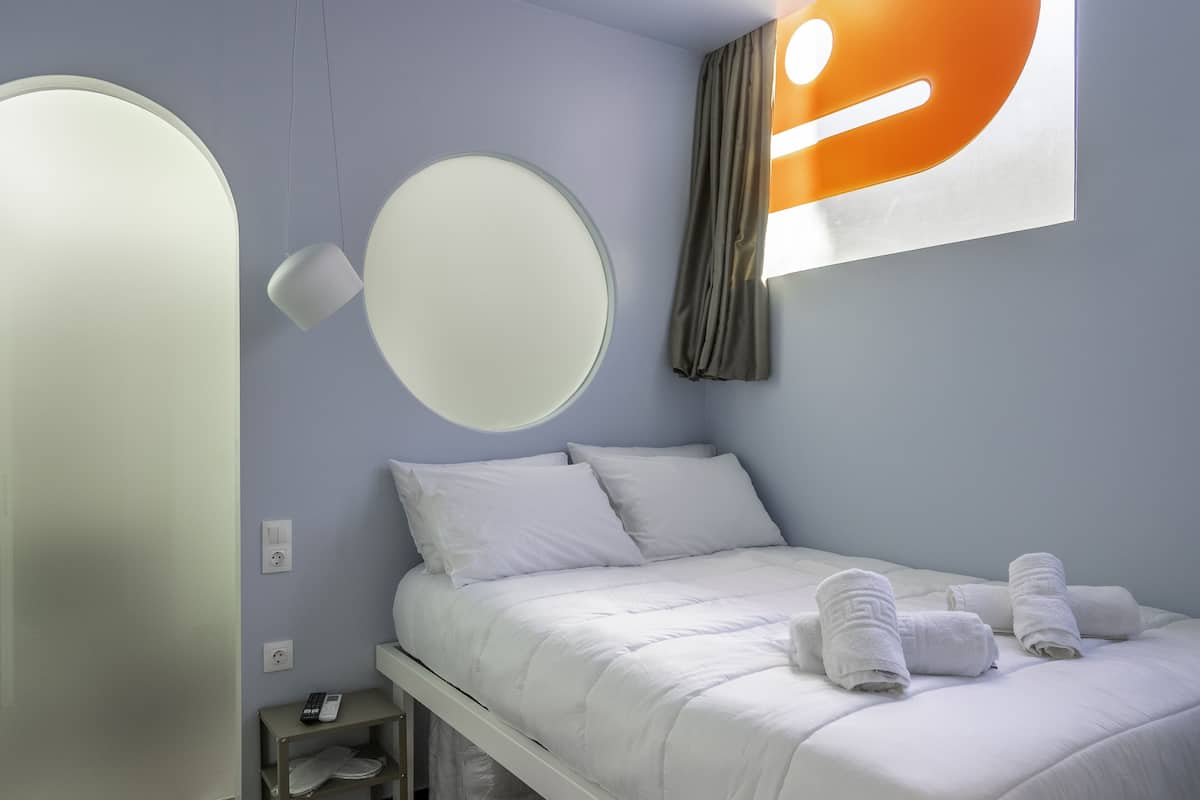 A neatly made bed with white bedding and towels, a bedside table with a remote control, and a wall-mounted round mirror.