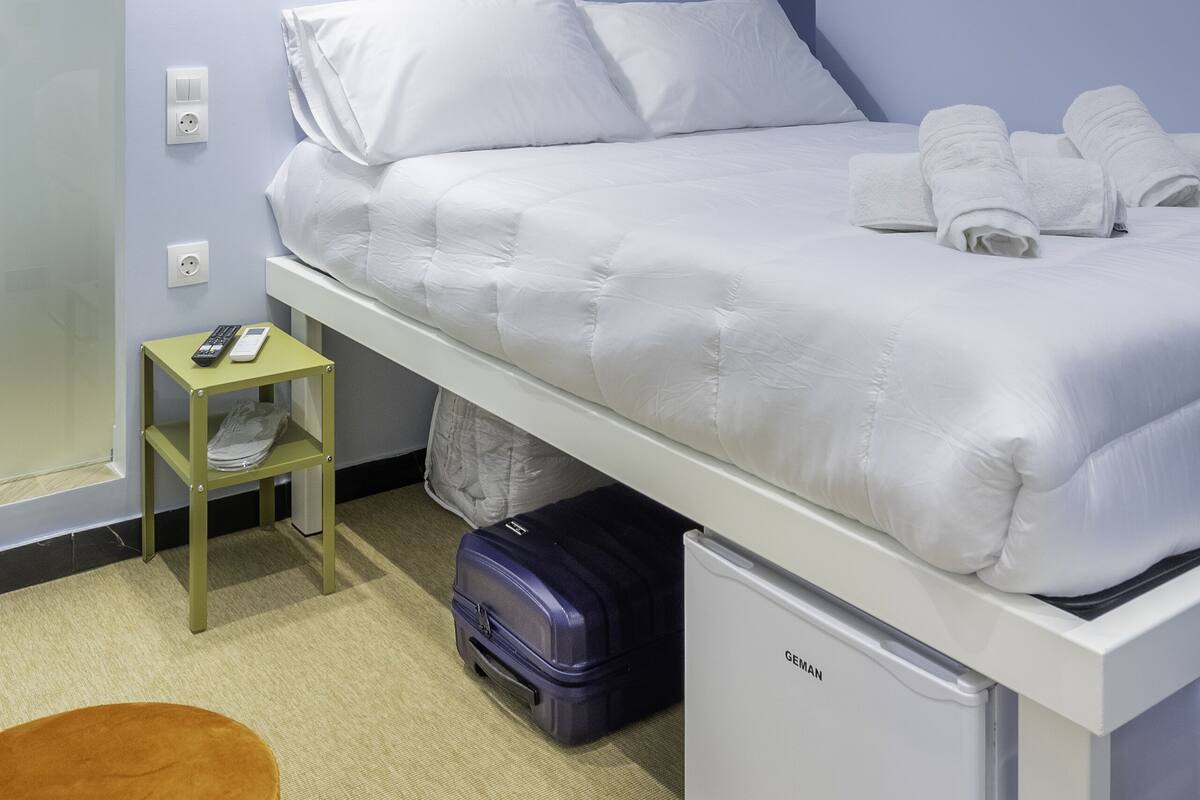 A modern hotel room with a neatly made bed, a small bedside table with a remote control, a suitcase, and a compact refrigerator.