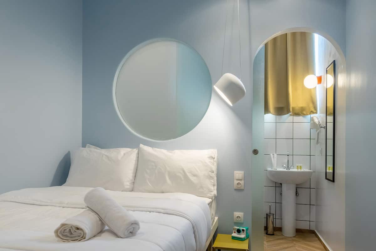 A bedroom with a bed, a round mirror, a bedside lamp, a small table, and a bathroom with a sink and mirror.
