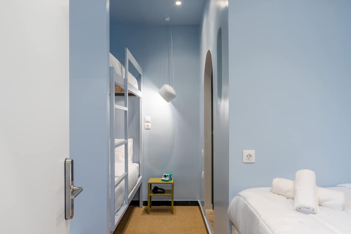 A hotel room with a bunk bed, a small table with a cup and a hat, a bed with rolled towels, and a light fixture.