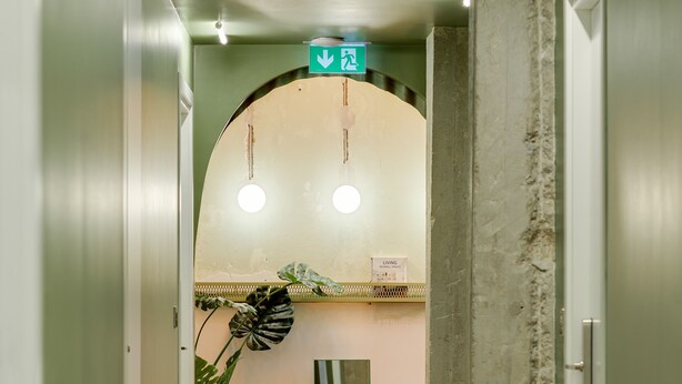 A hallway with a green exit sign, a plant, and a mirror on the wall. - Cabins Sleeping Mets (Athens)
