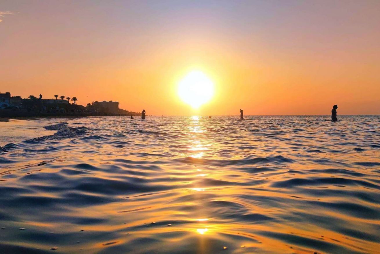 A beach scene with the sun setting over the water, silhouettes of people, and a distant shoreline with buildings.