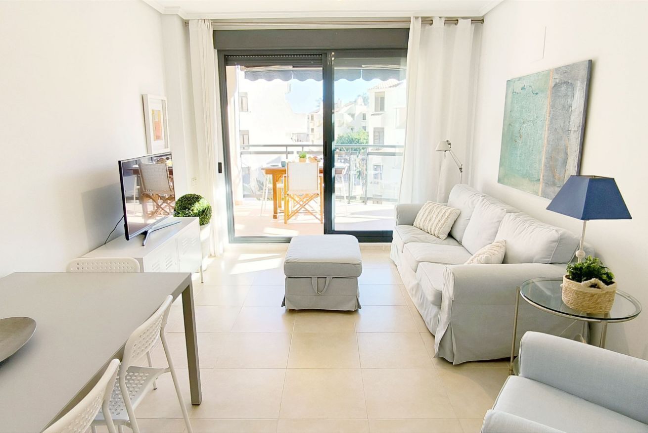 A modern living room with a sofa, a glass table, a TV, and a balcony with a table and chairs.