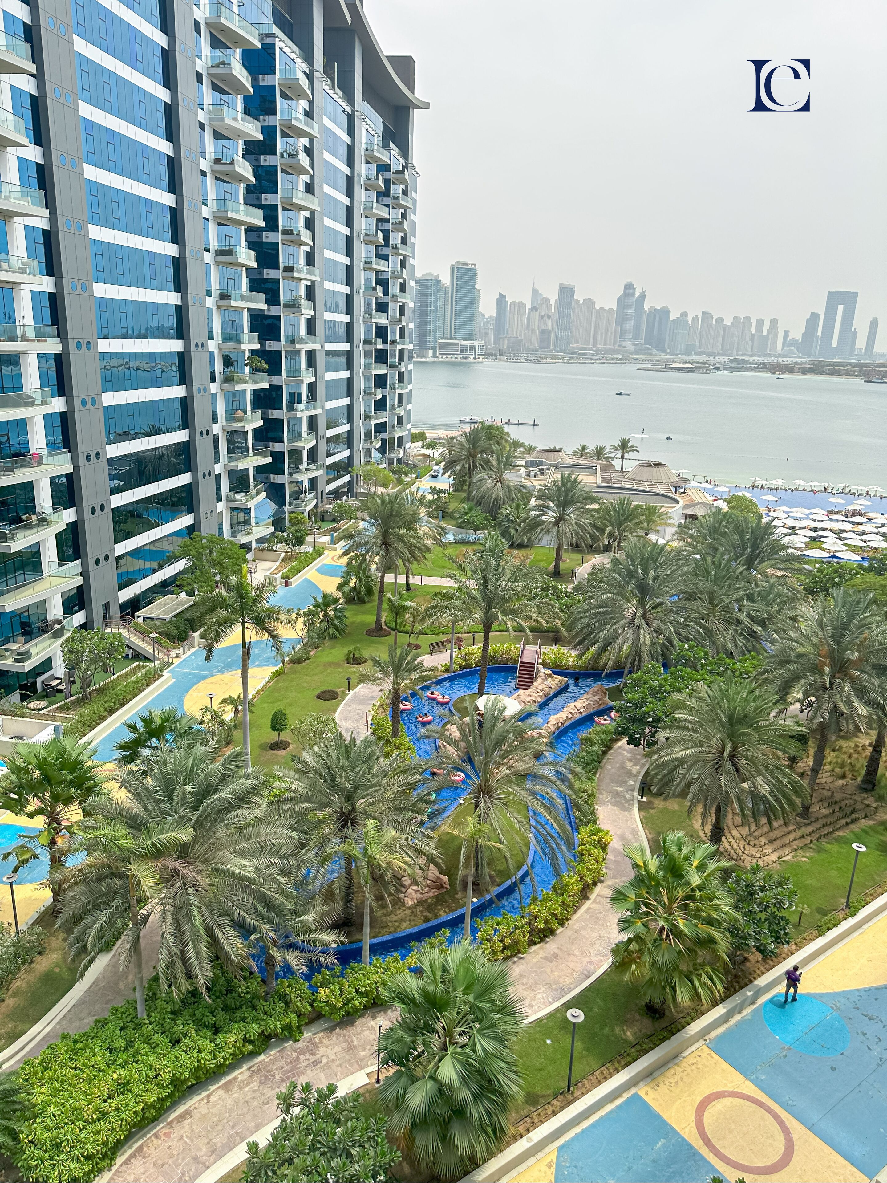 Un immeuble résidentiel de grande hauteur doté de balcons, d’un espace piscine agrémenté de palmiers et offrant une vue sur les gratte-ciel d’une ville, au-delà d’une étendue d’eau.