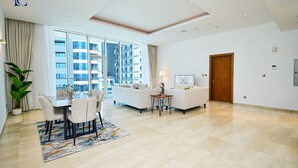 A modern hotel room with a dining area, a sofa, a lamp, and a view of the cityscape. - Premium 3BR Partial Sea View OCEANA (Dubai)