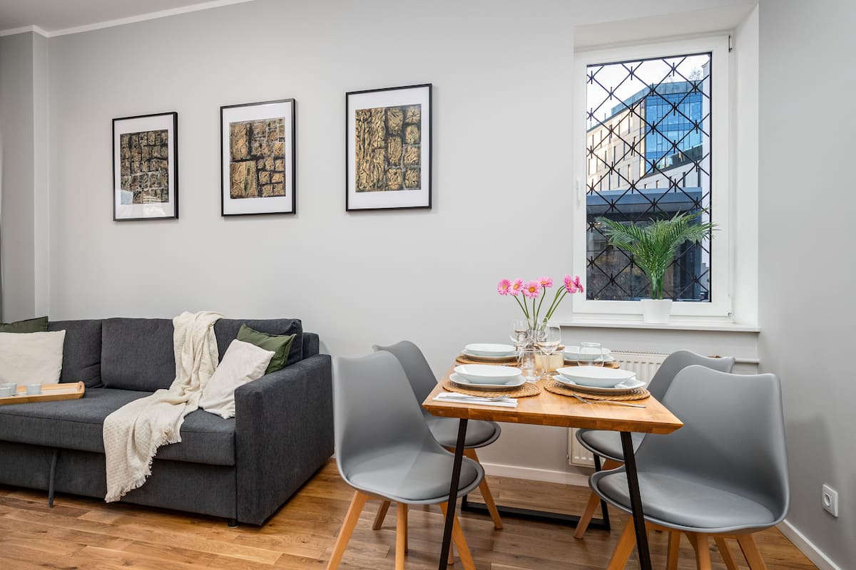 A modern living room with a grey sofa, a wooden dining table set with plates and glasses, and three framed artworks on the wall.
