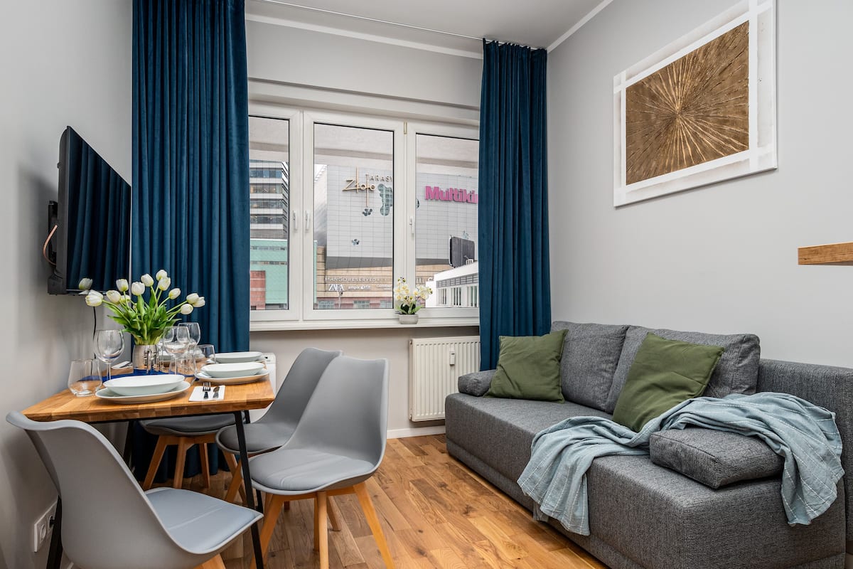 A modern living room with a grey sofa, a dining table with chairs, and a window with curtains.
