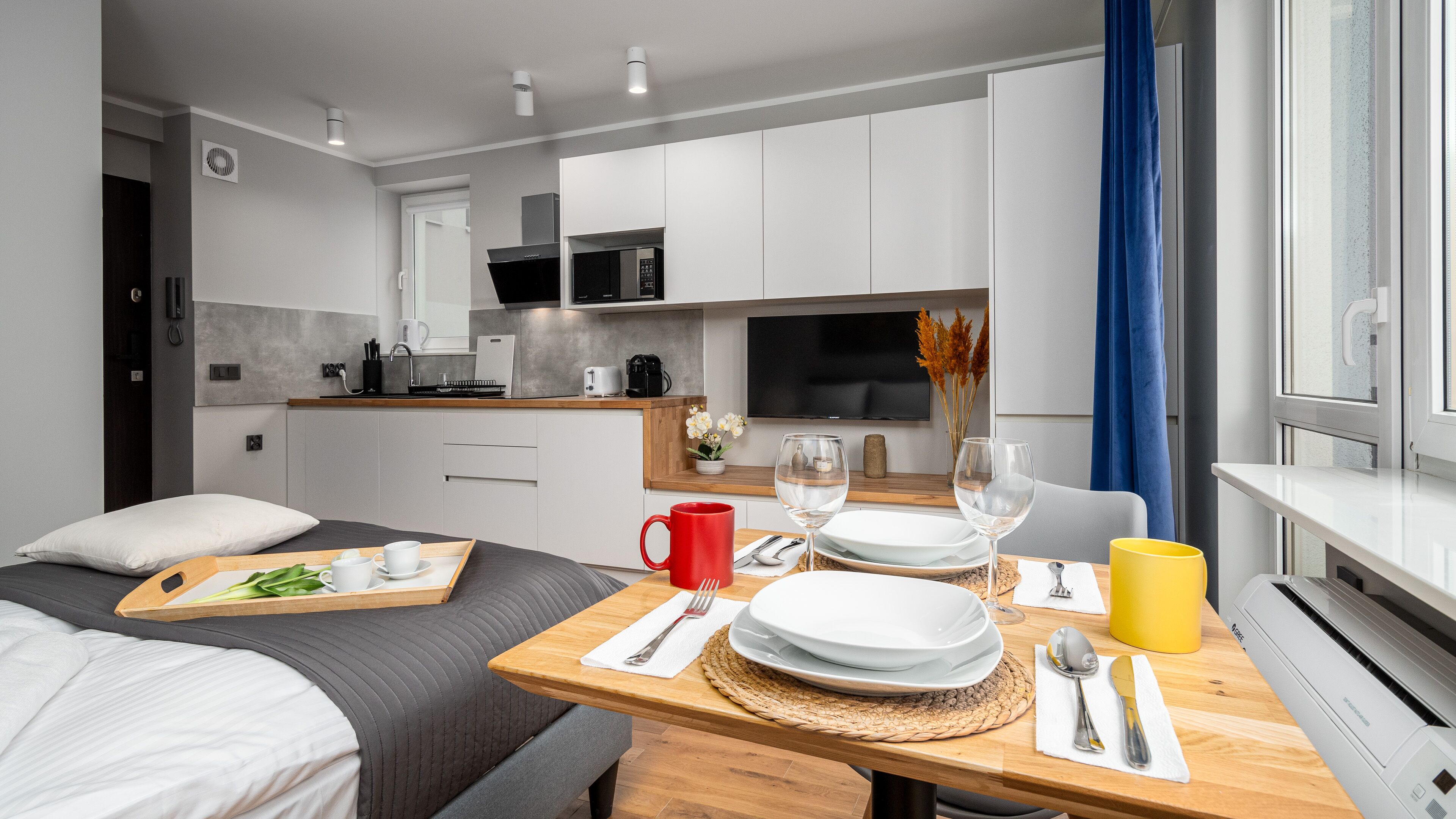 A modern hotel room with a dining area, kitchenette, and a large window with curtains.