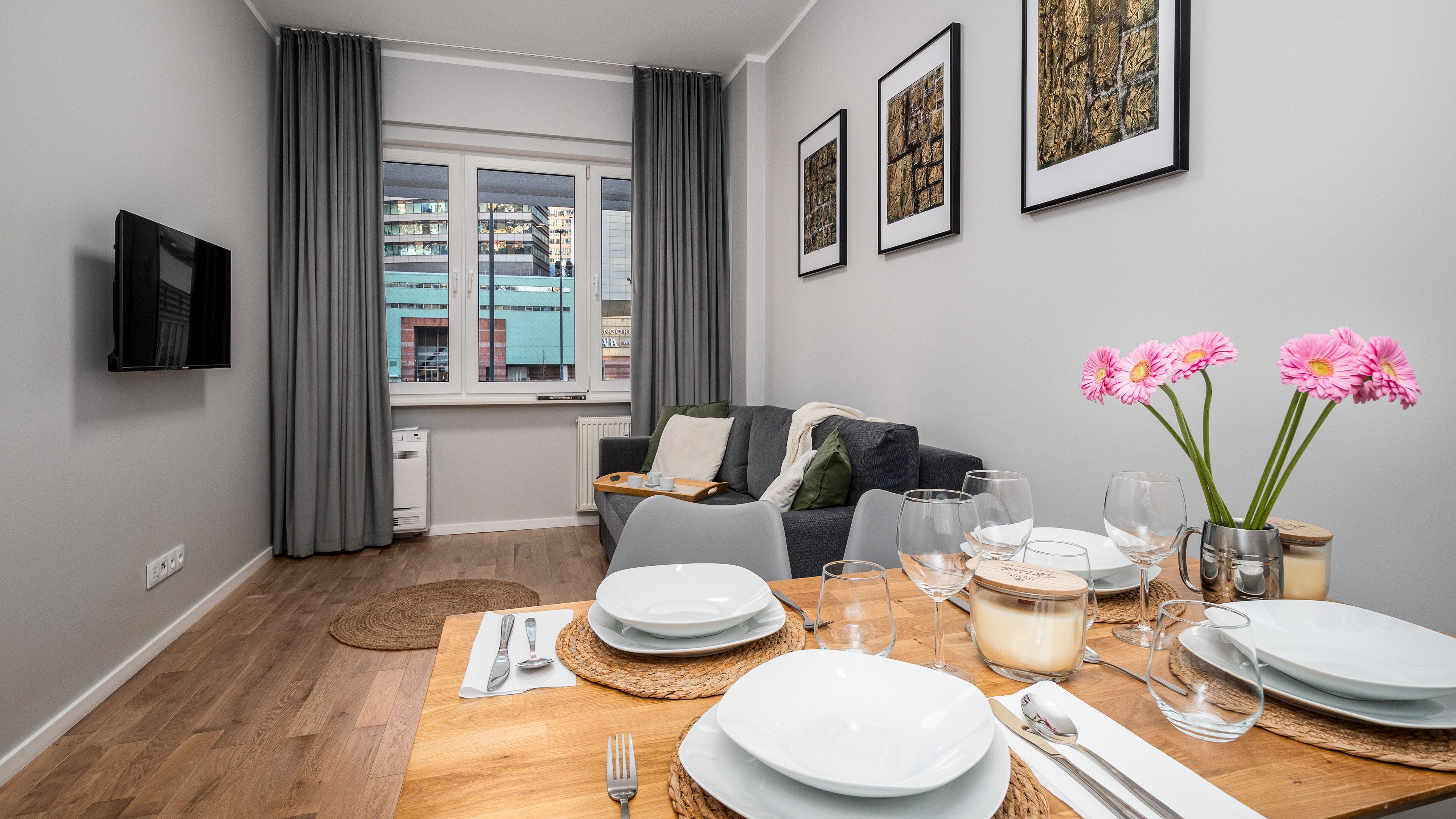 A modern living room with a dining table set for four, a sofa, a TV mounted on the wall, and a window with curtains.