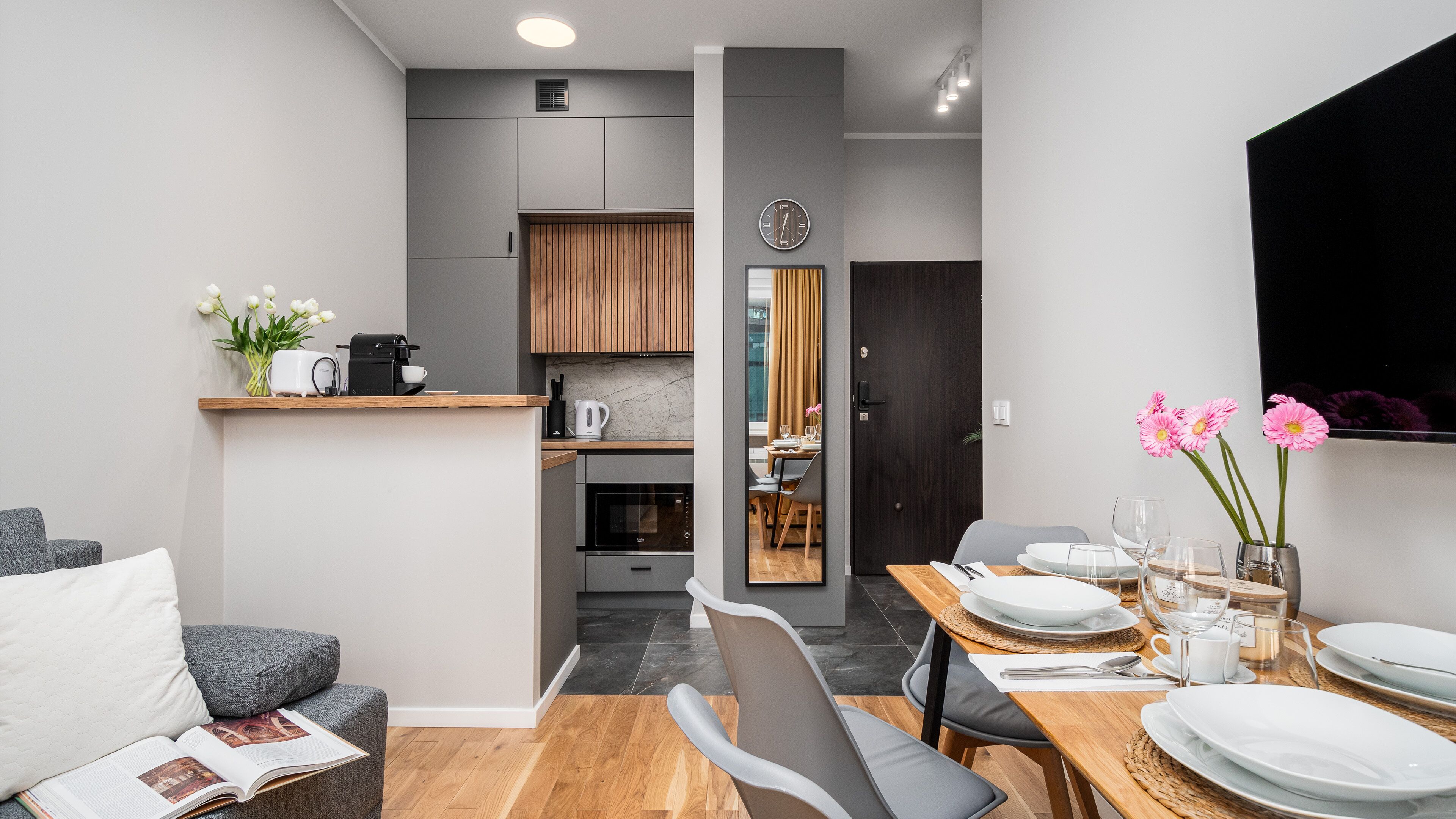 A modern kitchen with grey cabinets, a built-in oven, and a dining area with a wooden table set for two.
