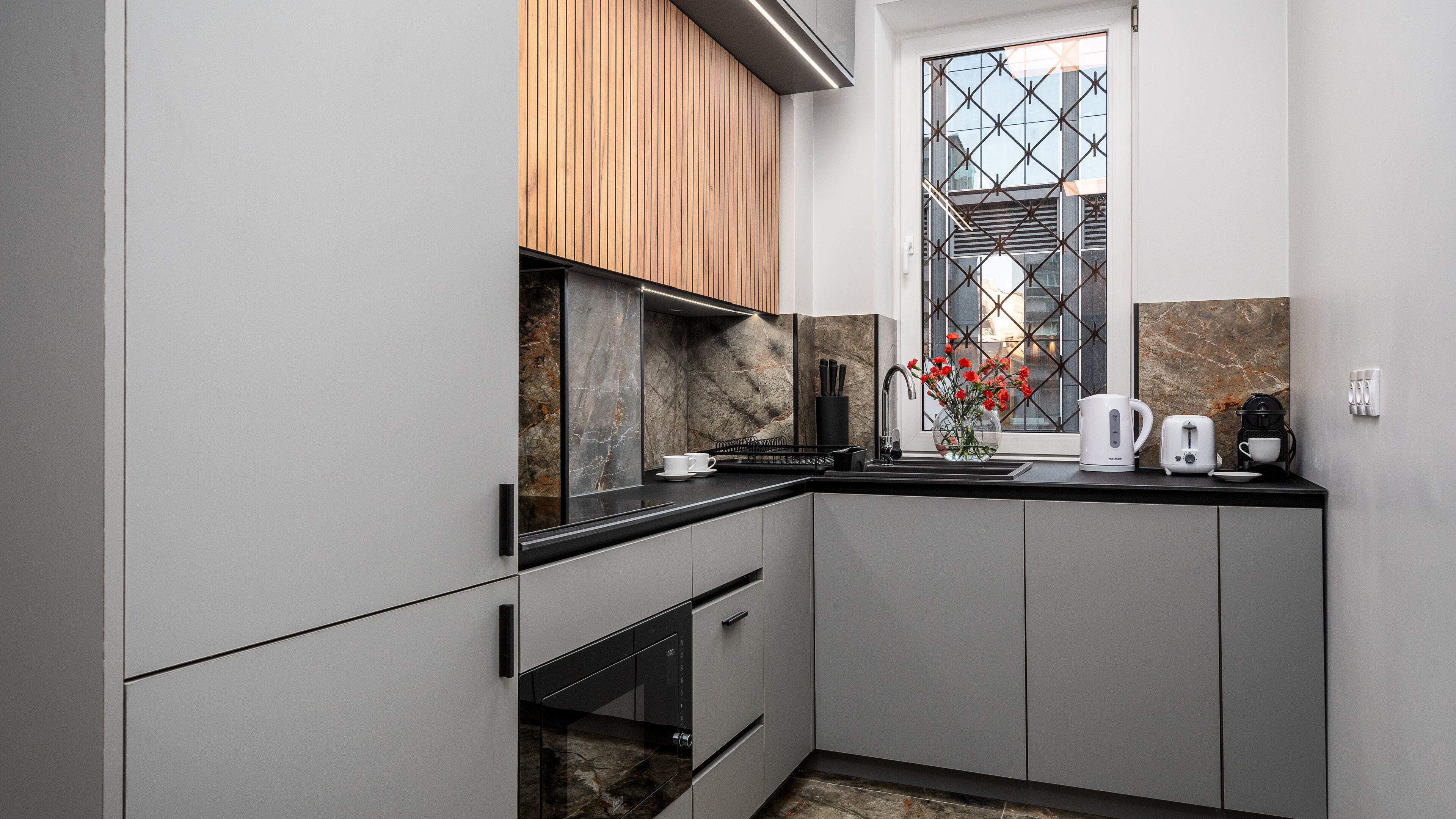 Une cuisine moderne avec des meubles de couleur grise, un élément supérieur en bois, un pare-feu en marbre et une fenêtre ornée d’une grille métallique.