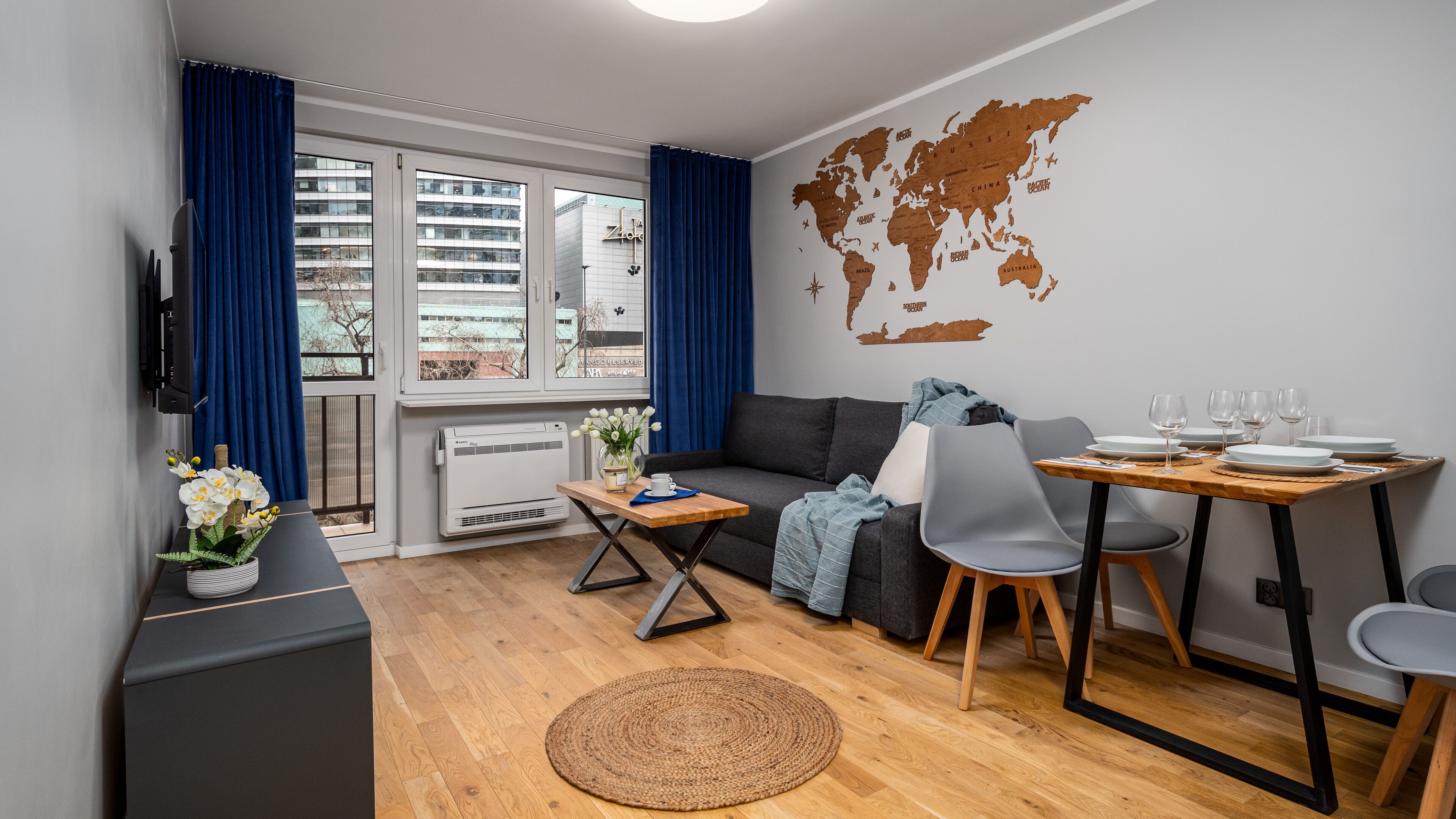 Ein modernes Wohnzimmer mit einer Couch, einem Esstisch und Stühlen. An der Wand hängt eine Weltkarte, ein Fernsehgerät und ein Fenster mit Vorhängen.
