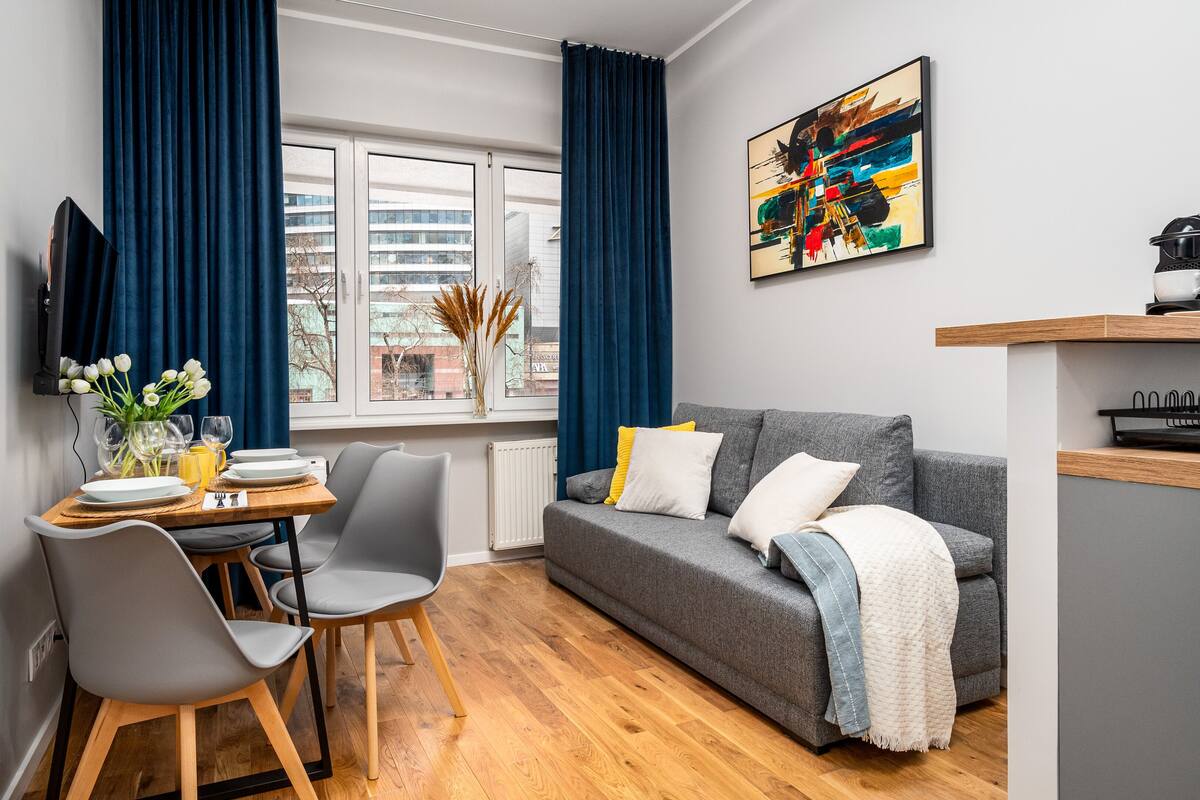 A modern living room with a grey sofa, a dining table with chairs, and a large window with blue curtains.