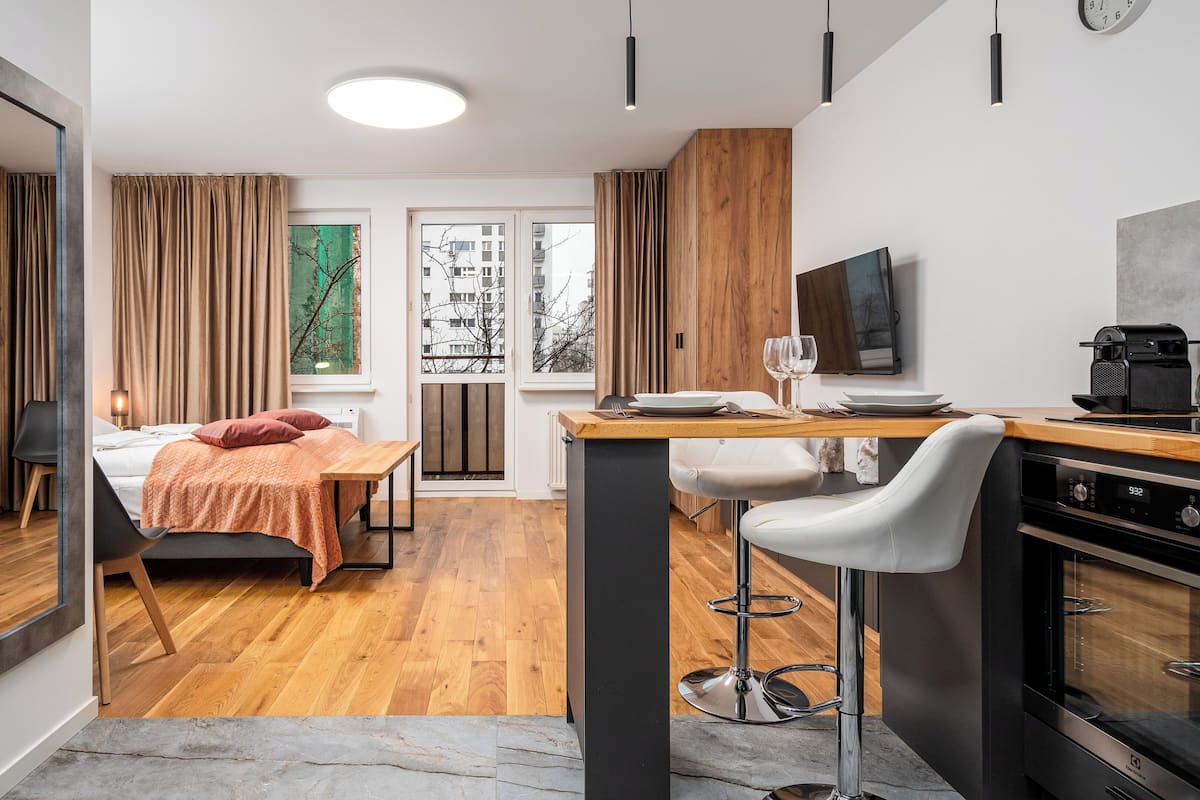 A modern hotel room with a wooden floor, a bed with a red throw, a dining area with a bar, and a TV mounted on the wall.