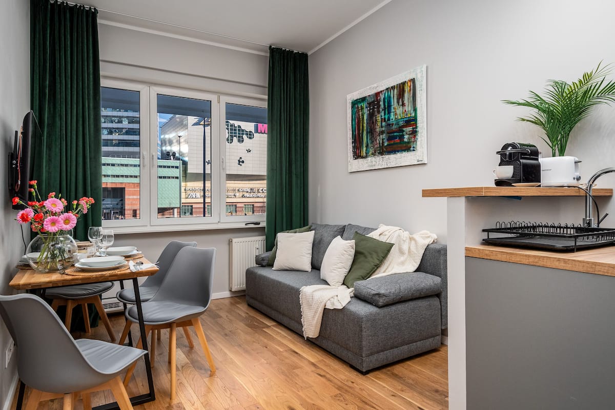 A compact living space with a dining area, a grey sofa, a small kitchen area, and a view of a building through a window.