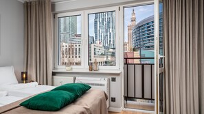 Et moderne hotelværelse med et stort vindue, der giver udsigt over byen, en seng med grønne puder og et natbord med en lampe.