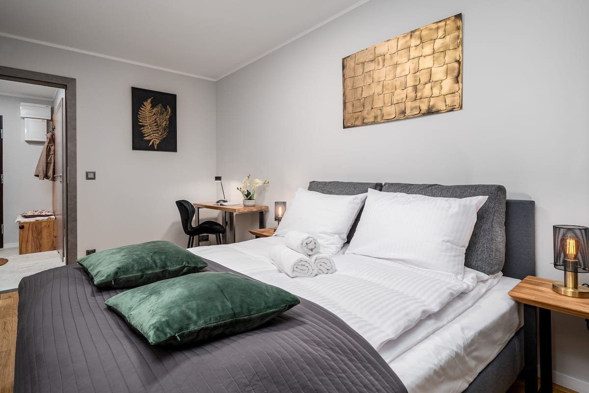 A modern bedroom with a large bed, green pillows, a desk with a chair, a small table with flowers, and a wall art piece.