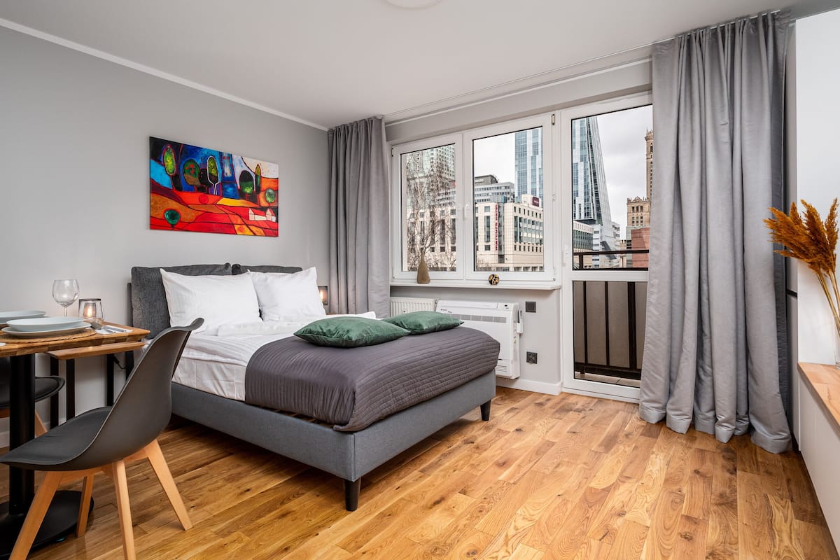 A modern hotel room with a bed, a desk with a chair, a window with a city view, and a painting on the wall.