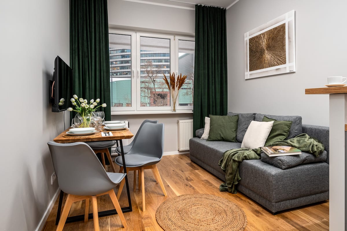 A modern living room with a grey sofa, a dining table with chairs, a TV mounted on the wall, and a window with curtains.