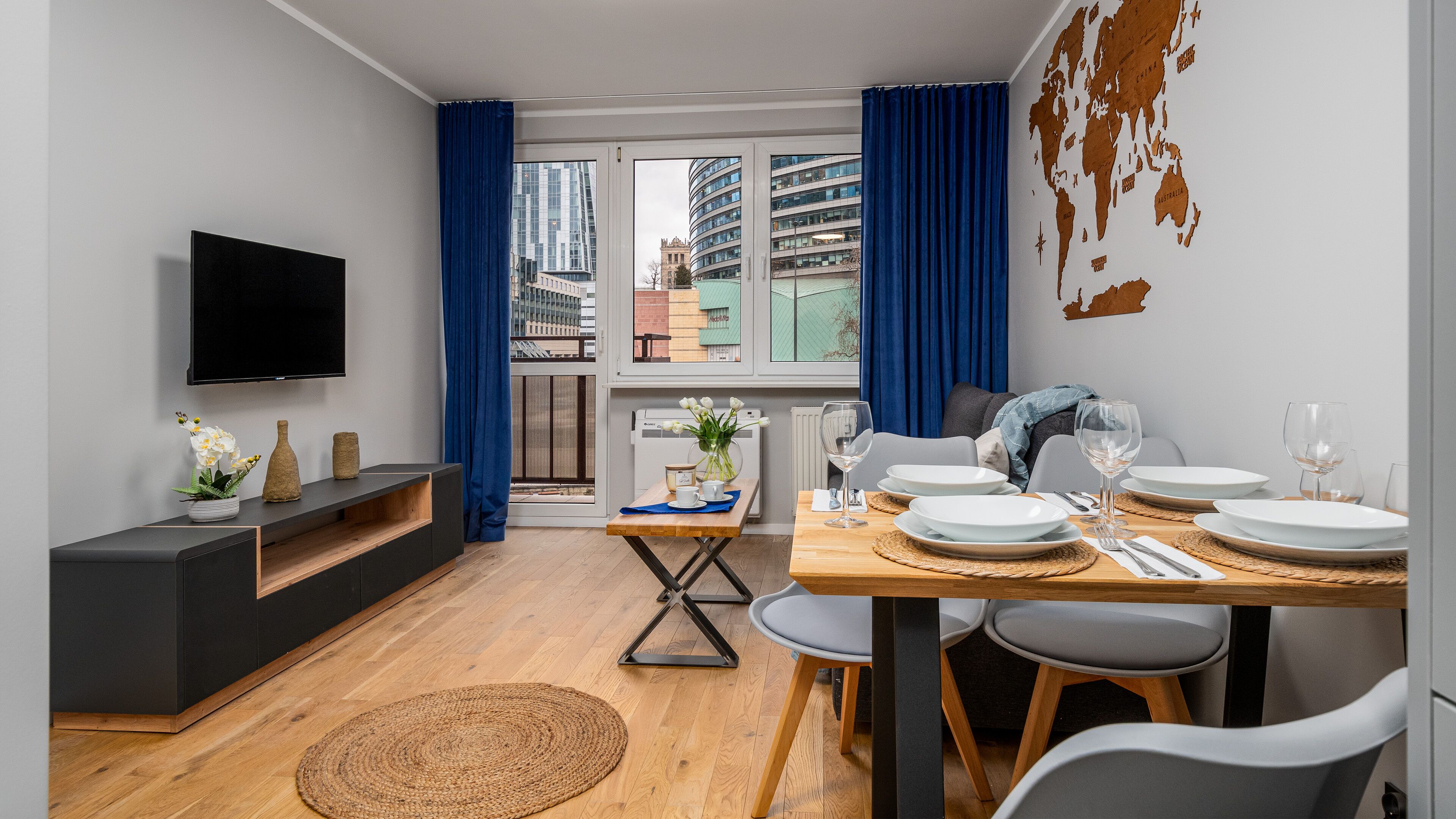 Een moderne eetruimte met een houten tafel gedekt voor vier personen, een aan de muur bevestigde televisie en een uitzicht op de stads skyline door grote ramen.