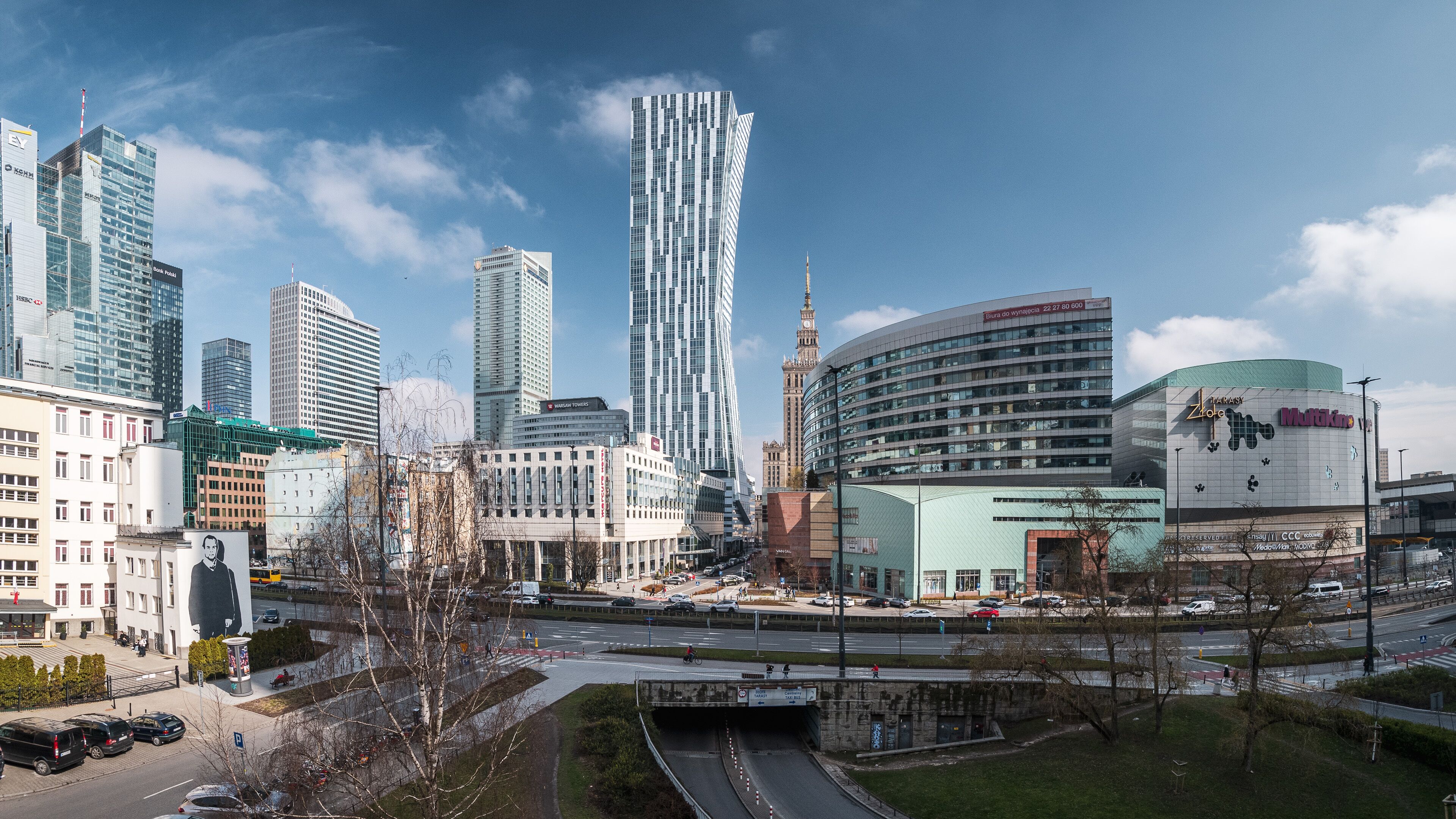 Paesaggio urbano con moderni grattacieli, una torre imponente e un edificio con facciata curva.
