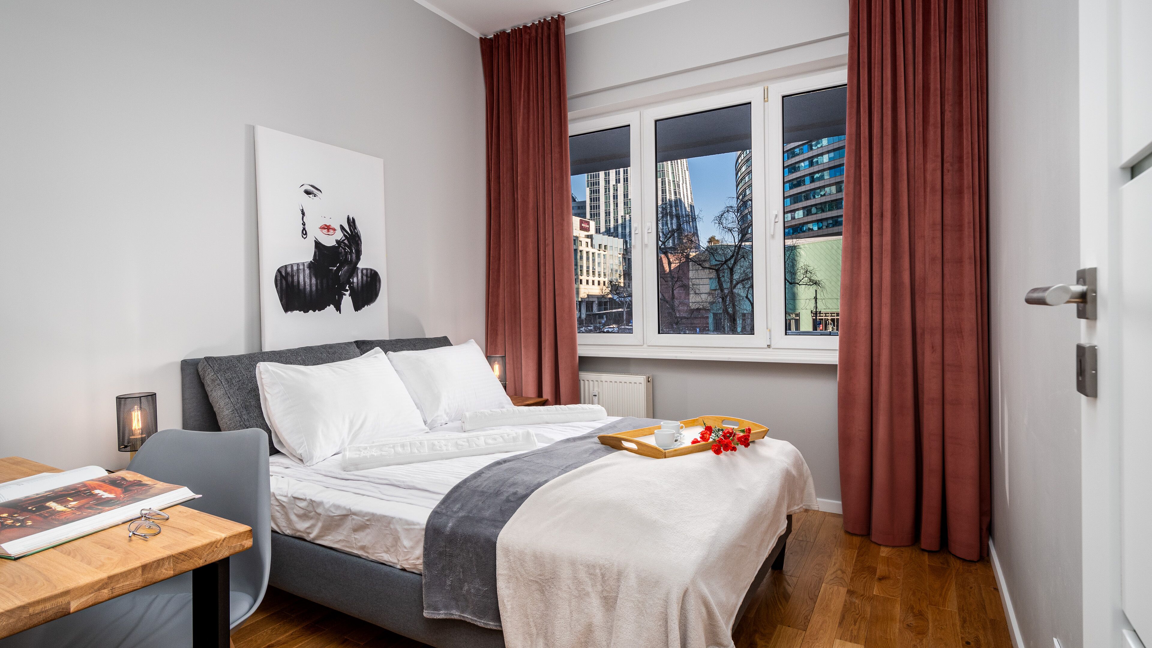 Una habitación de hotel moderna con cama, mesita de noche, ventana con cortinas, vista a la ciudad y un cuadro en la pared.