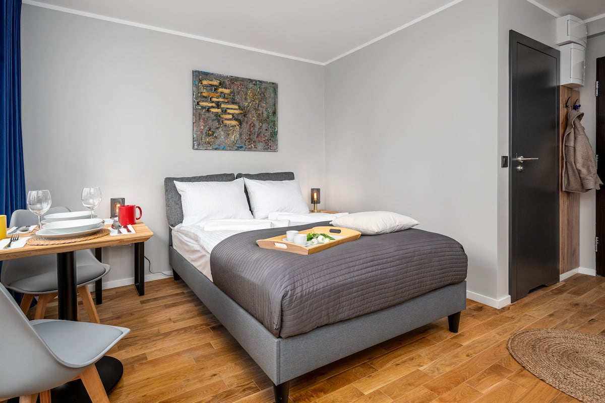 A modern hotel room with a bed, a desk with breakfast items, a chair, and a door.
