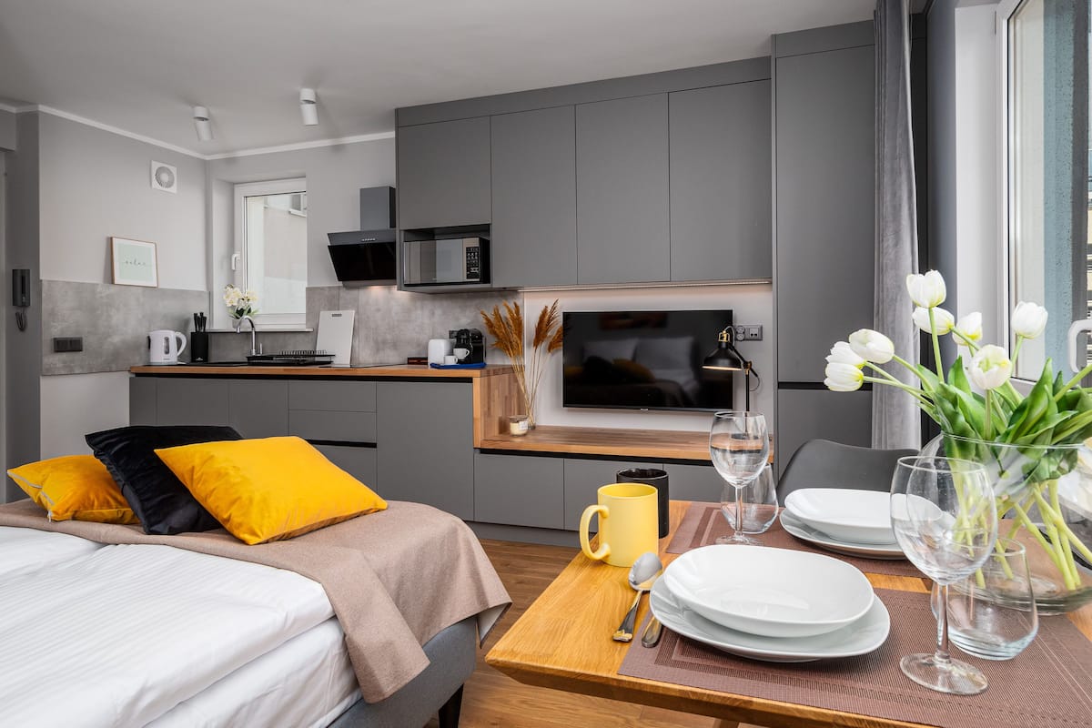 A modern hotel room with a bed, a dining table set for two, a TV, and a kitchen area.