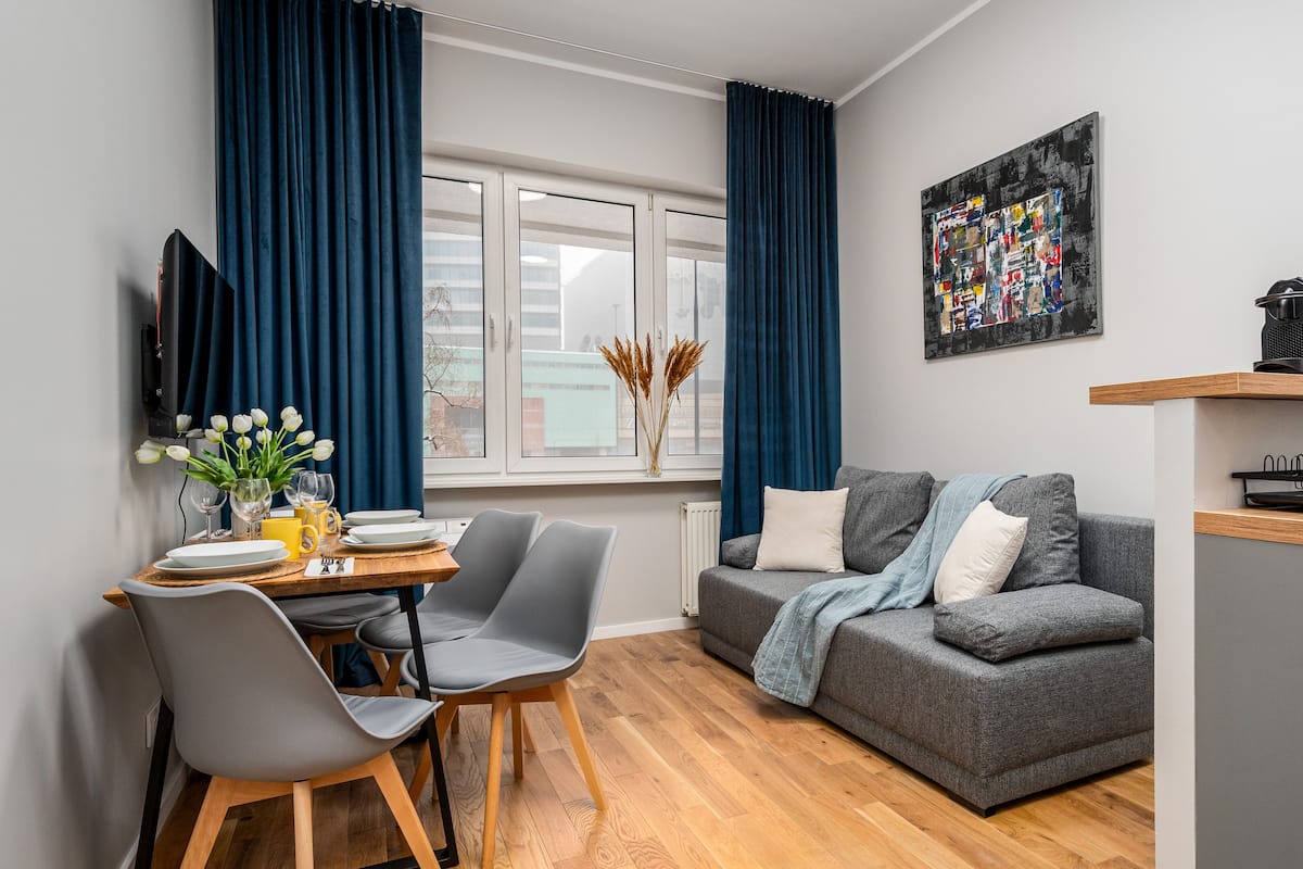 A modern living room with a grey sofa, a dining table with chairs, a TV mounted on the wall, and a window with blue curtains.