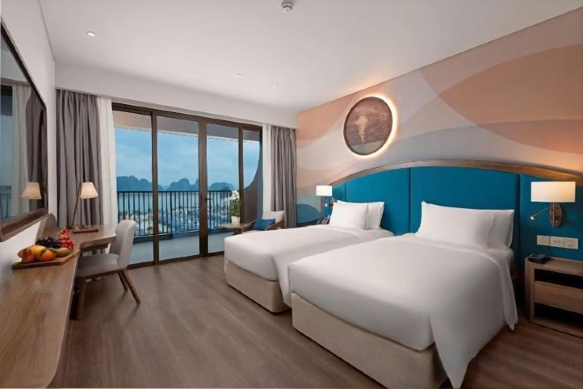 Ein Hotelzimmer mit zwei Betten, einem Schreibtisch mit Stuhl, einem Esstisch mit Früchten und einem großen Fenster mit Aussicht.