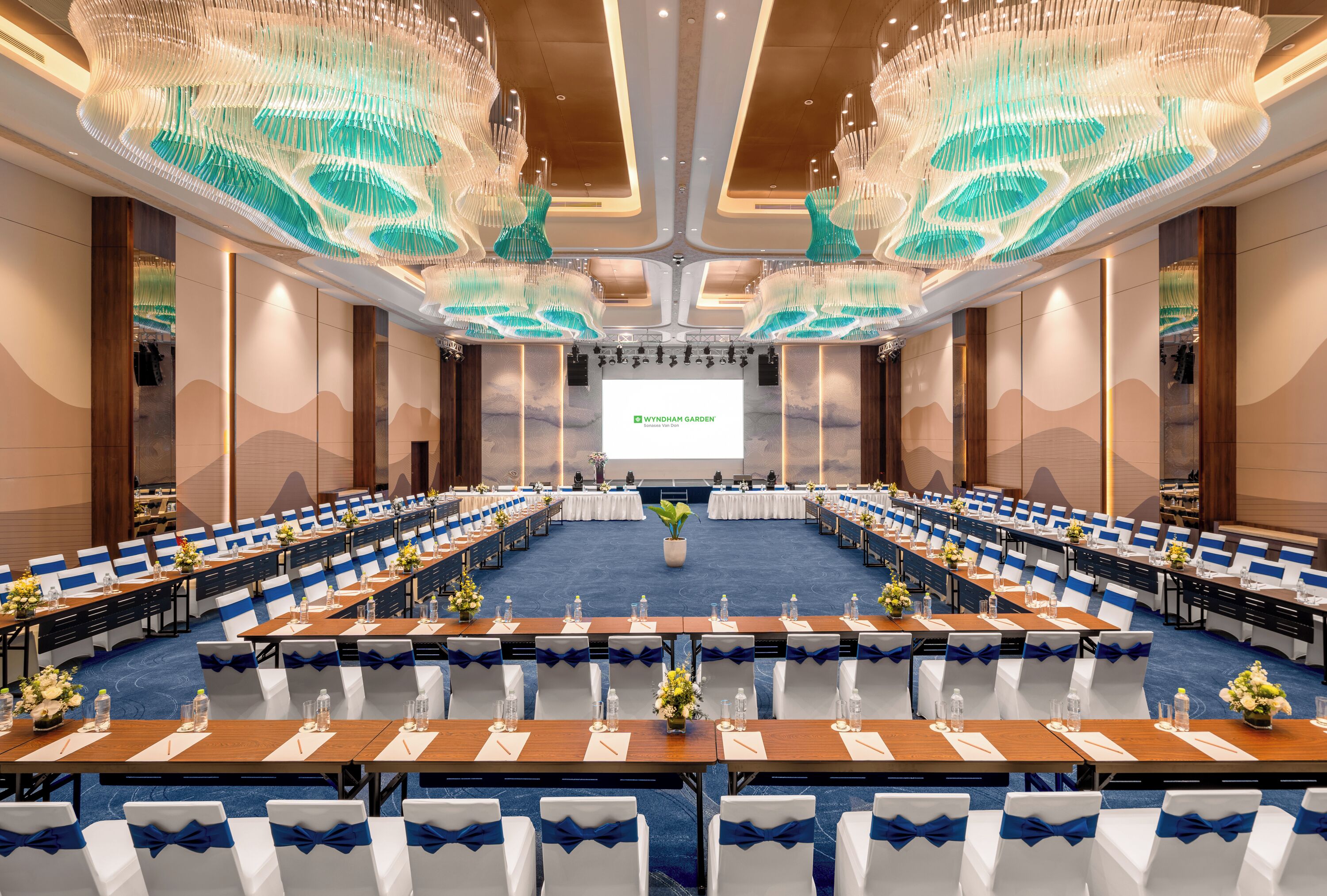 Une salle de conférence avec des rangées de tables et de chaises, un grand écran à l’avant et un lustre moderne.