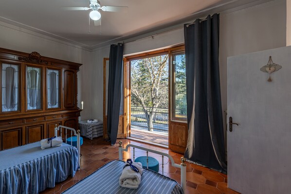A room with two beds, a large wooden cabinet, a ceiling fan, and a view of the outdoors through a large window.