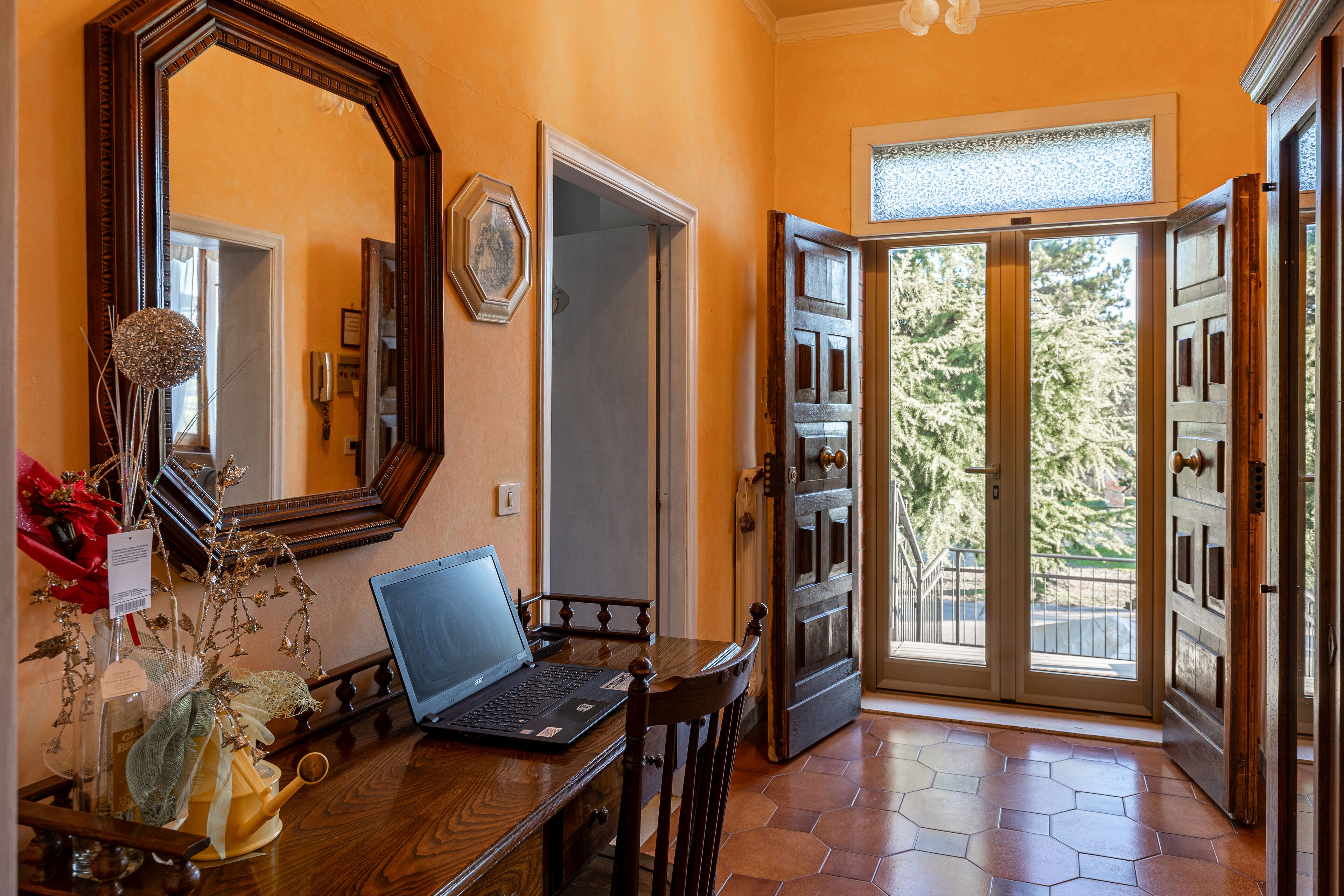 Una habitación con una mesa de madera, una computadora portátil, un espejo y una puerta que da a un balcón con vistas a árboles.