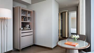 A modern hotel room with a minibar, a round dining table with a book, and a view of the hallway.