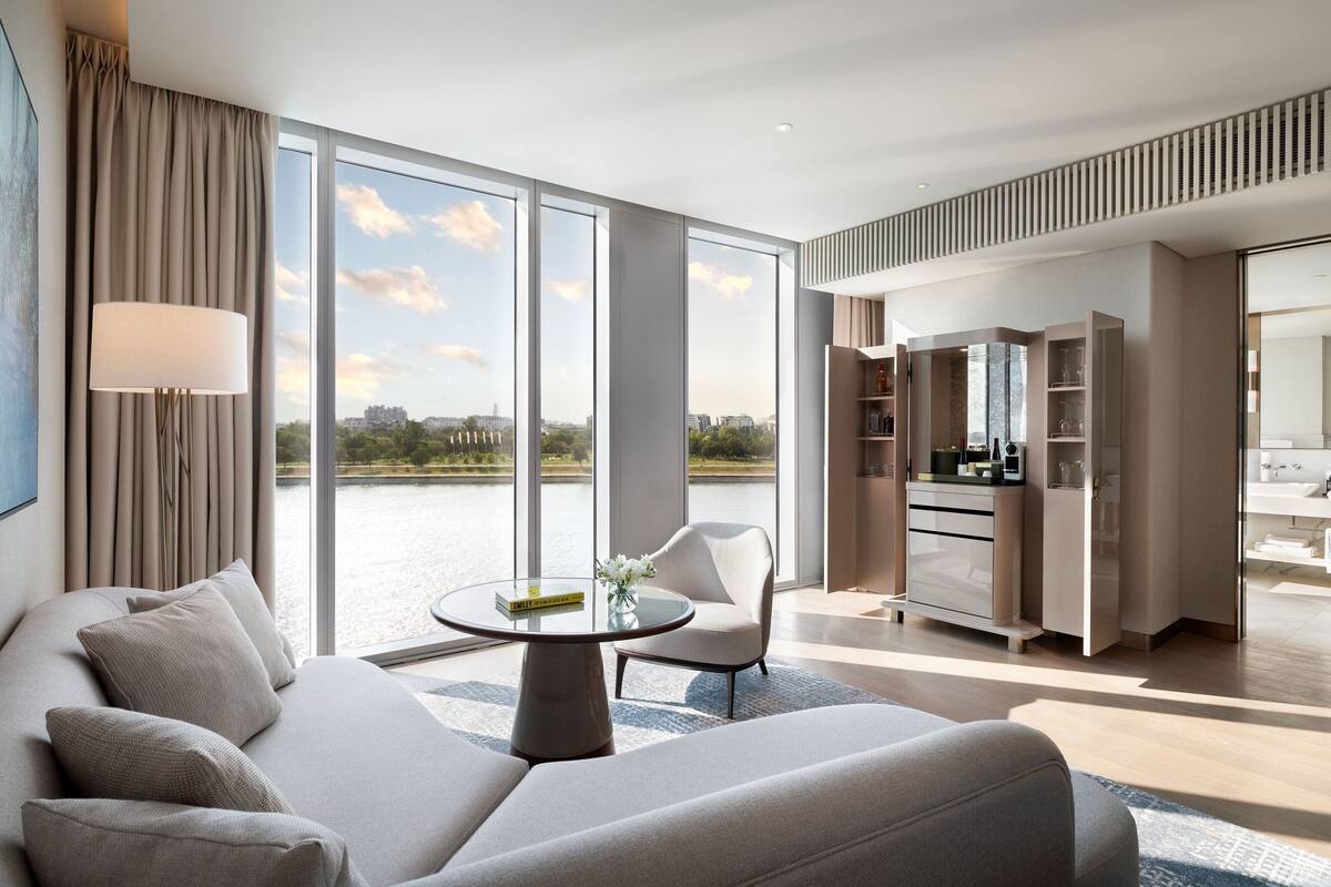 A modern hotel room with a large window, a sofa, a dining area, and a kitchen.