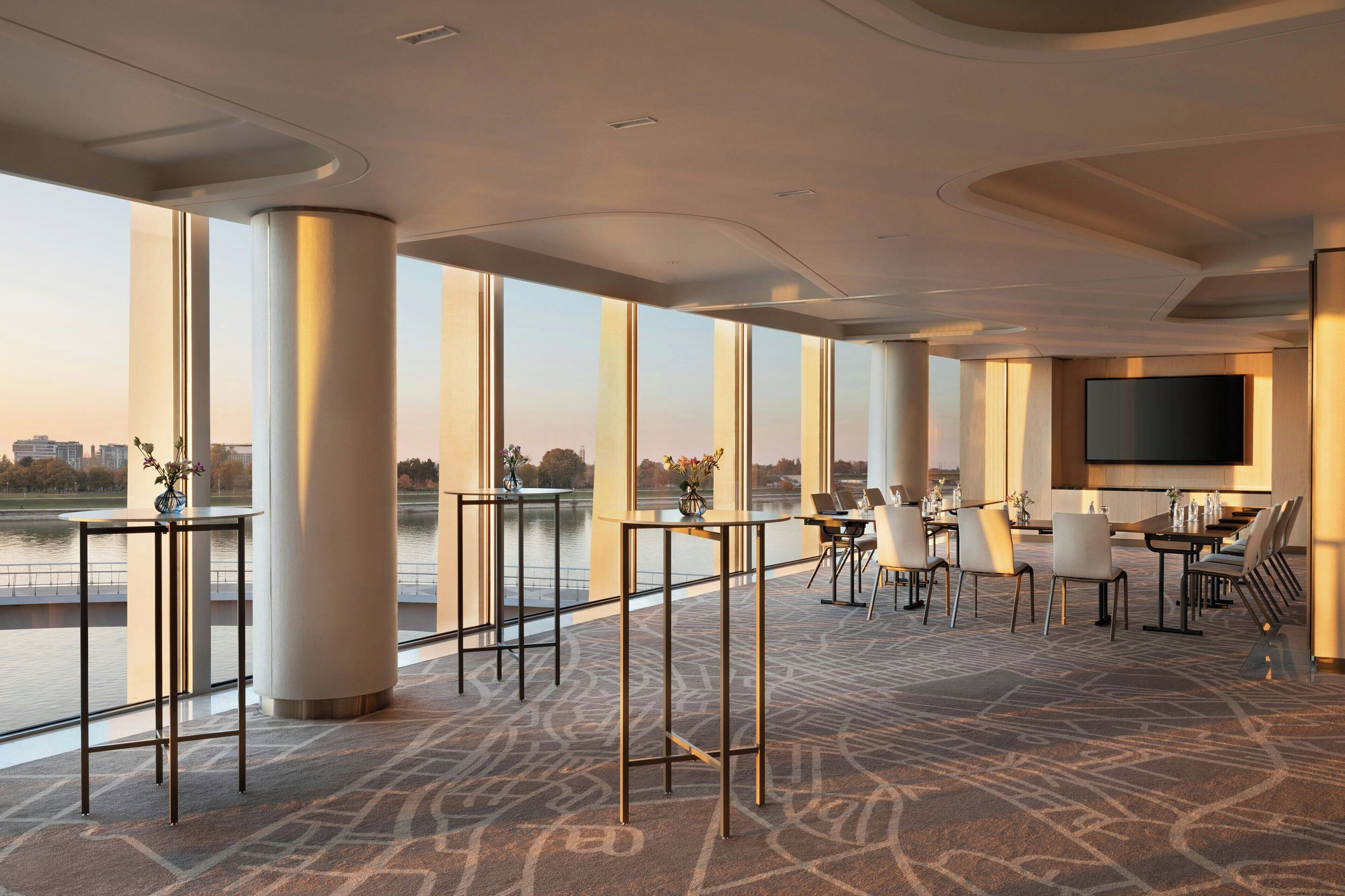 A spacious hotel lobby with large windows offering a view of a body of water, a flat-screen TV mounted on the wall, and a seating area with tables and chairs.