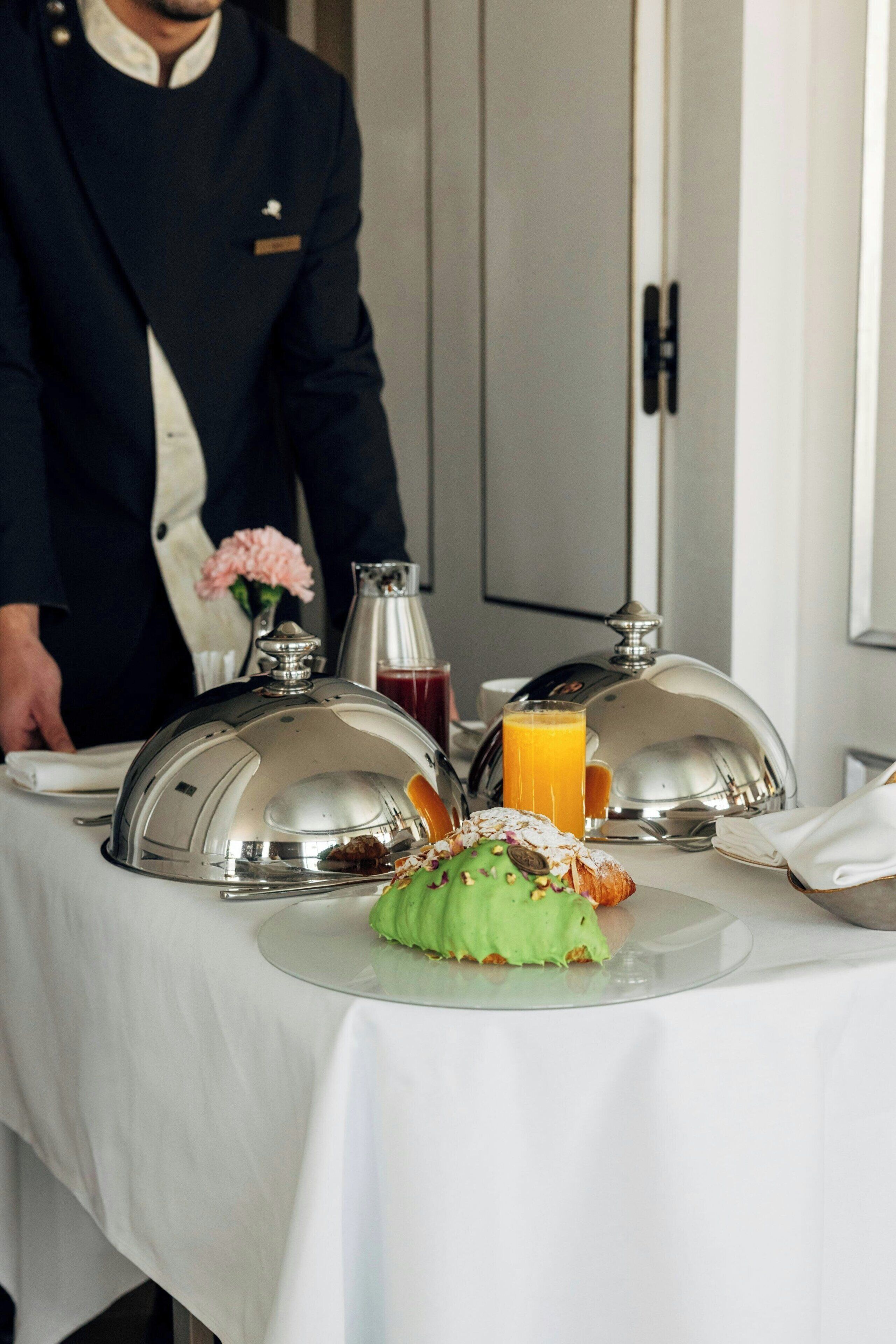 Ein Frühstücksstillleben mit einem silbernen Tablett mit Deckel, einem grünen, gezuckerten Gebäckstück, einem Glas Orangensaft und einer Kaffeekanne.