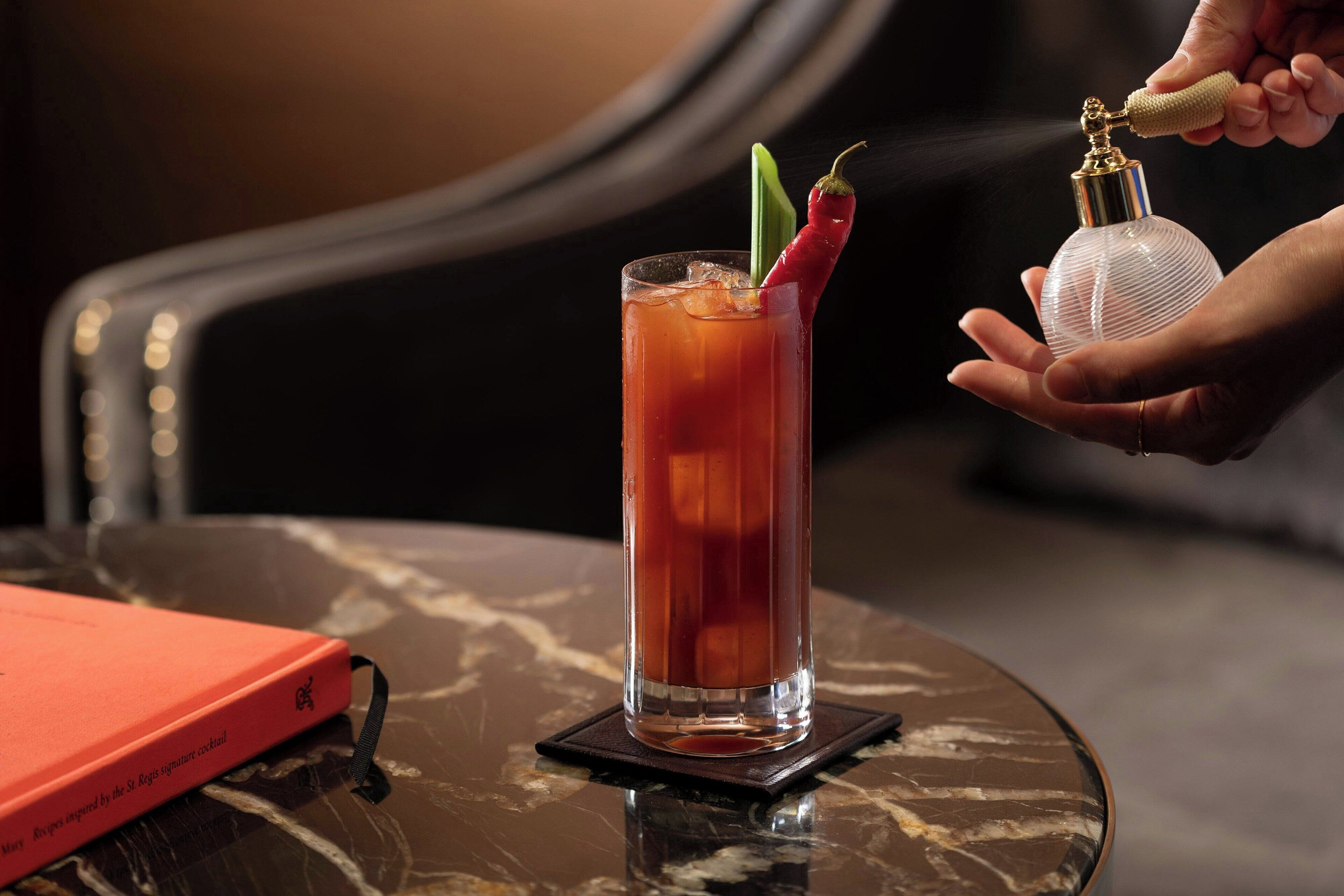 Un grand verre de Bloody Mary, garni d’un piment rouge et d’autres décorations, tenu dans des mains, posé sur une table en marbre à côté d’un livre.