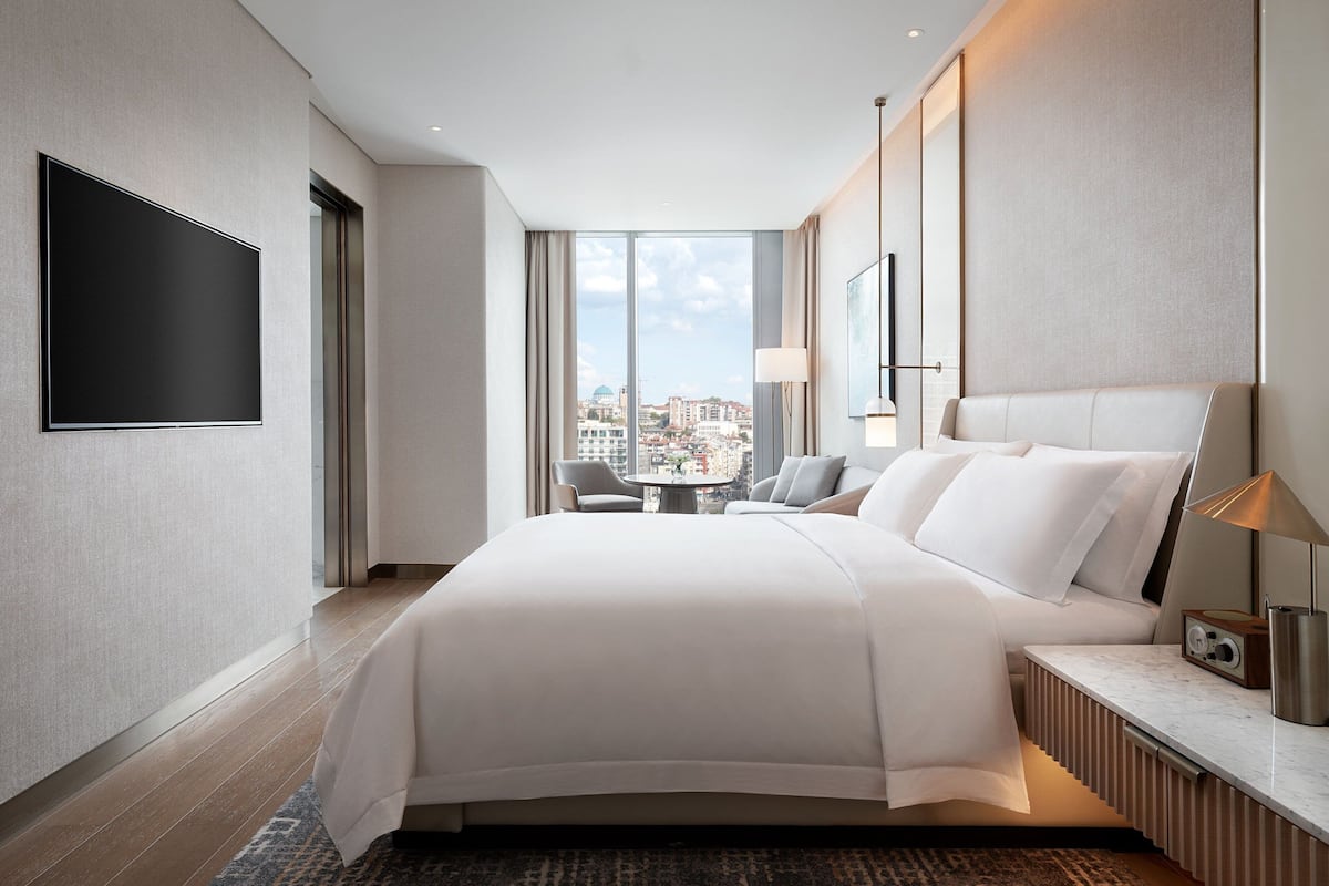 A modern hotel room with a large bed, a flat-screen TV, a desk with a lamp, and a view of the city through large windows.