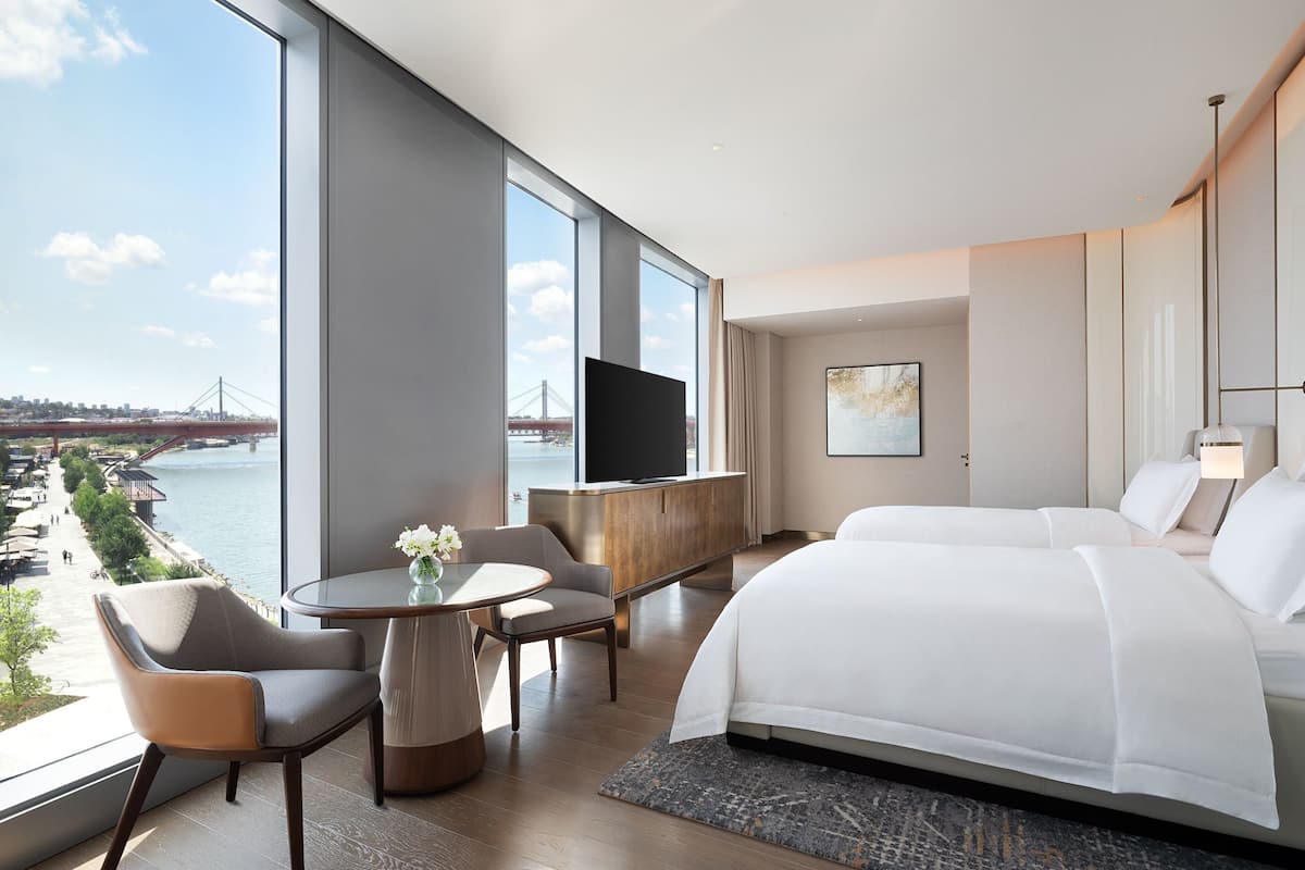 A modern hotel room with a large window offering a view of a bridge and river, a bed, a TV, a dining area with a table and chairs, and a small desk.