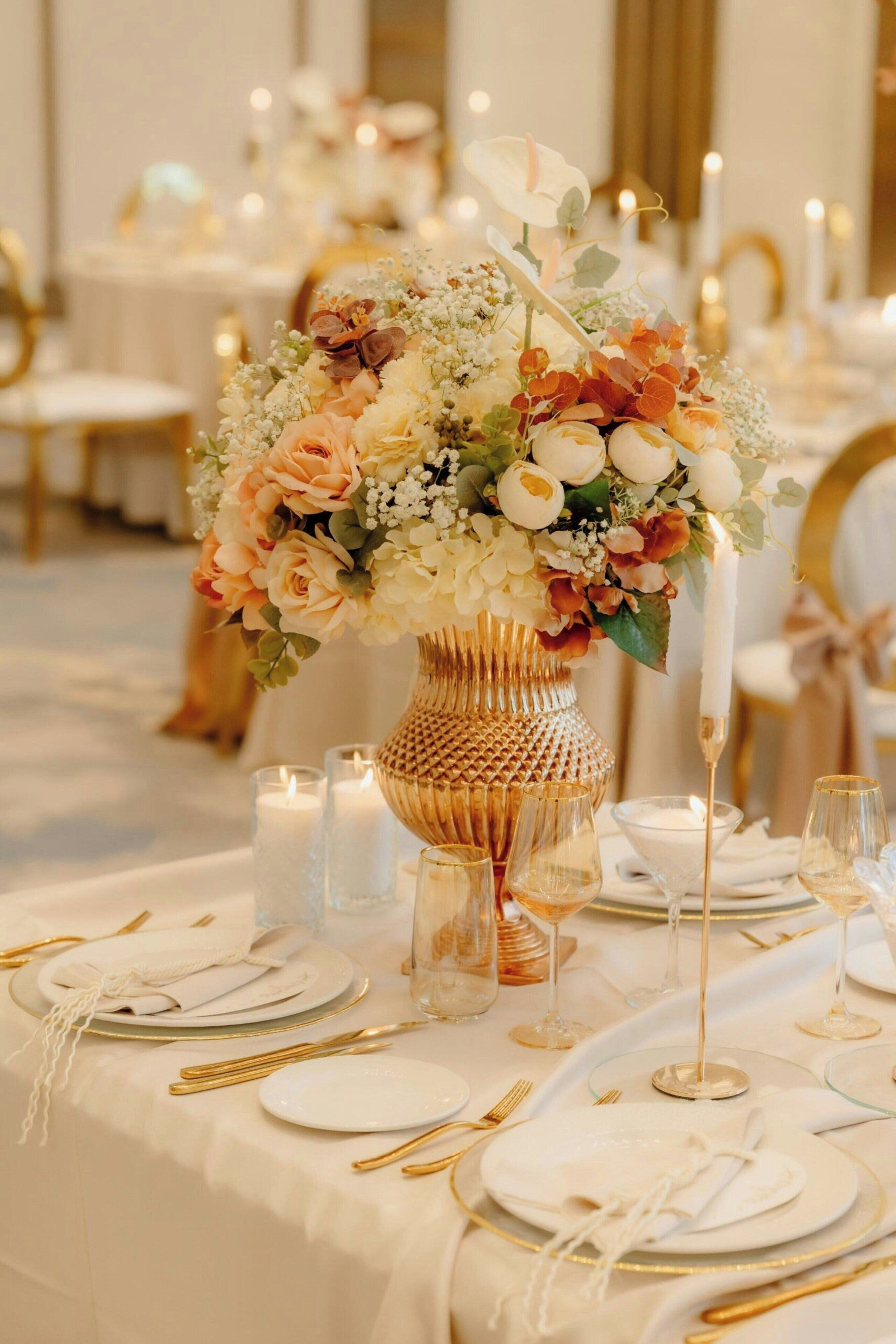 Una mesa dispuesta con platos blancos, cubiertos dorados y un centro de mesa de flores en un florero decorativo.