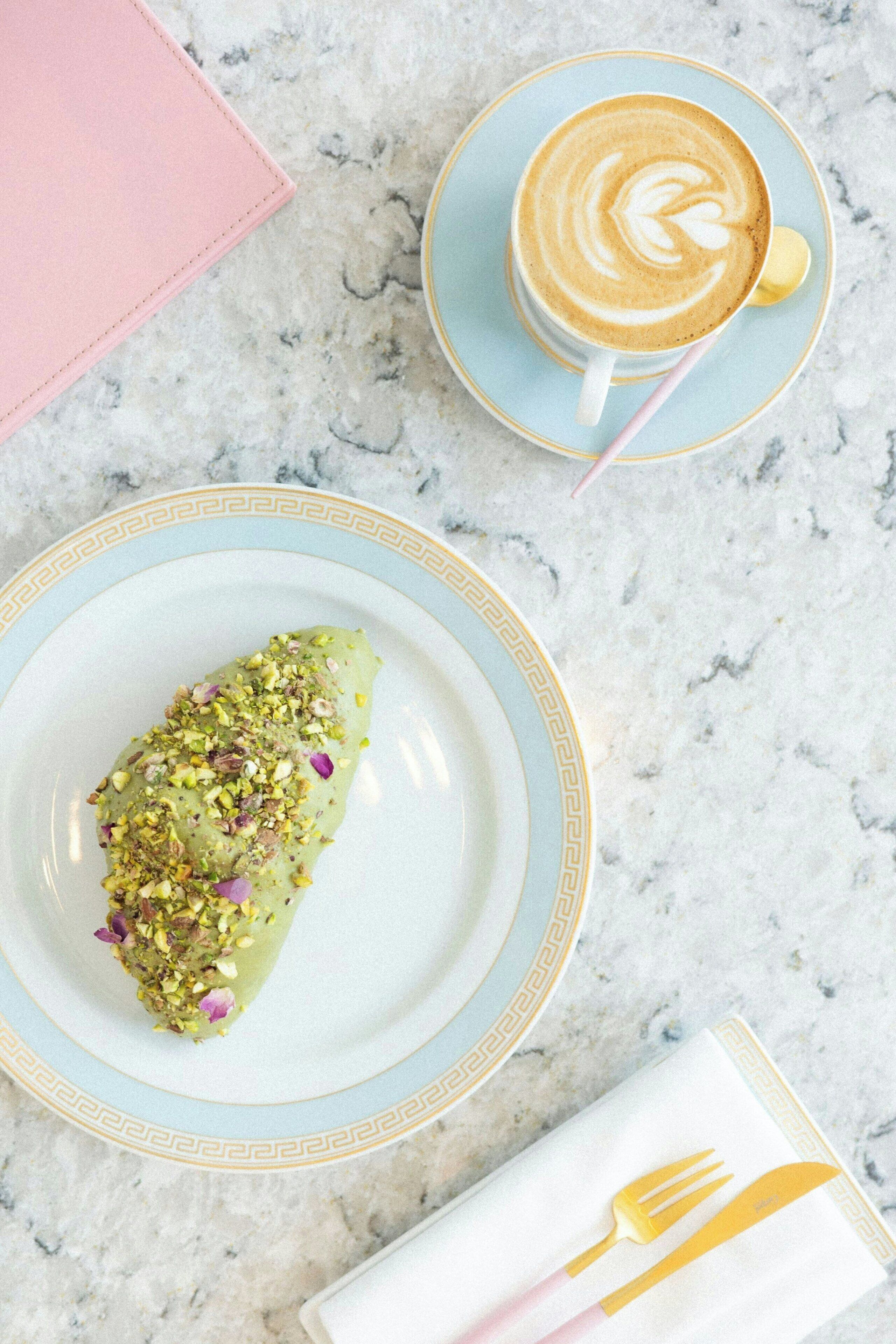 A dessert on a plate, a cup of coffee, and a napkin with cutlery on a marble surface.
