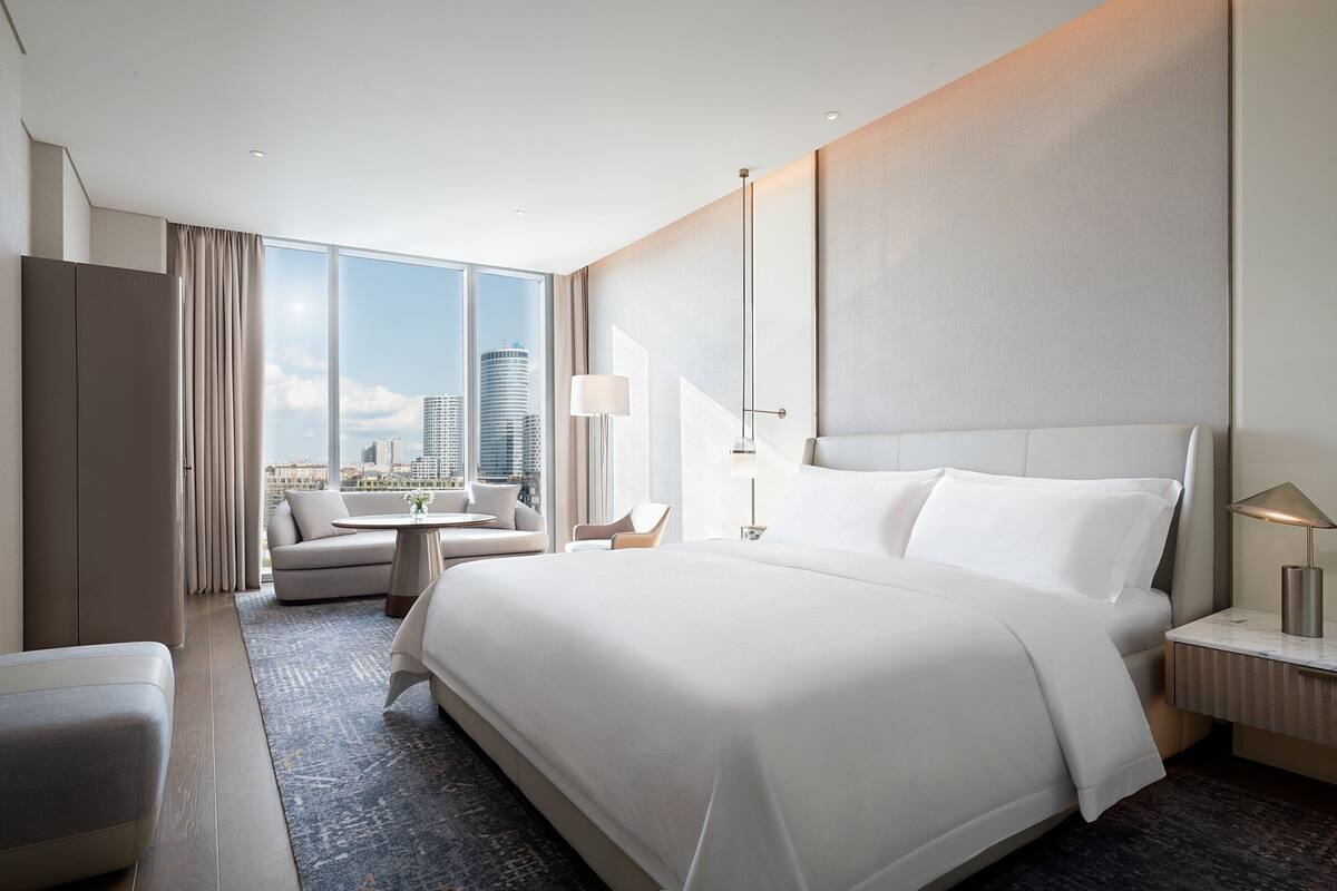 A modern hotel room with a large bed, a sofa, a desk, and a view of a cityscape.