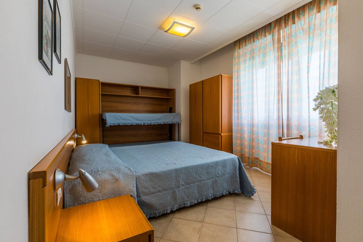 Un dormitorio con cama, mesita de noche, armario y ventana con cortinas.