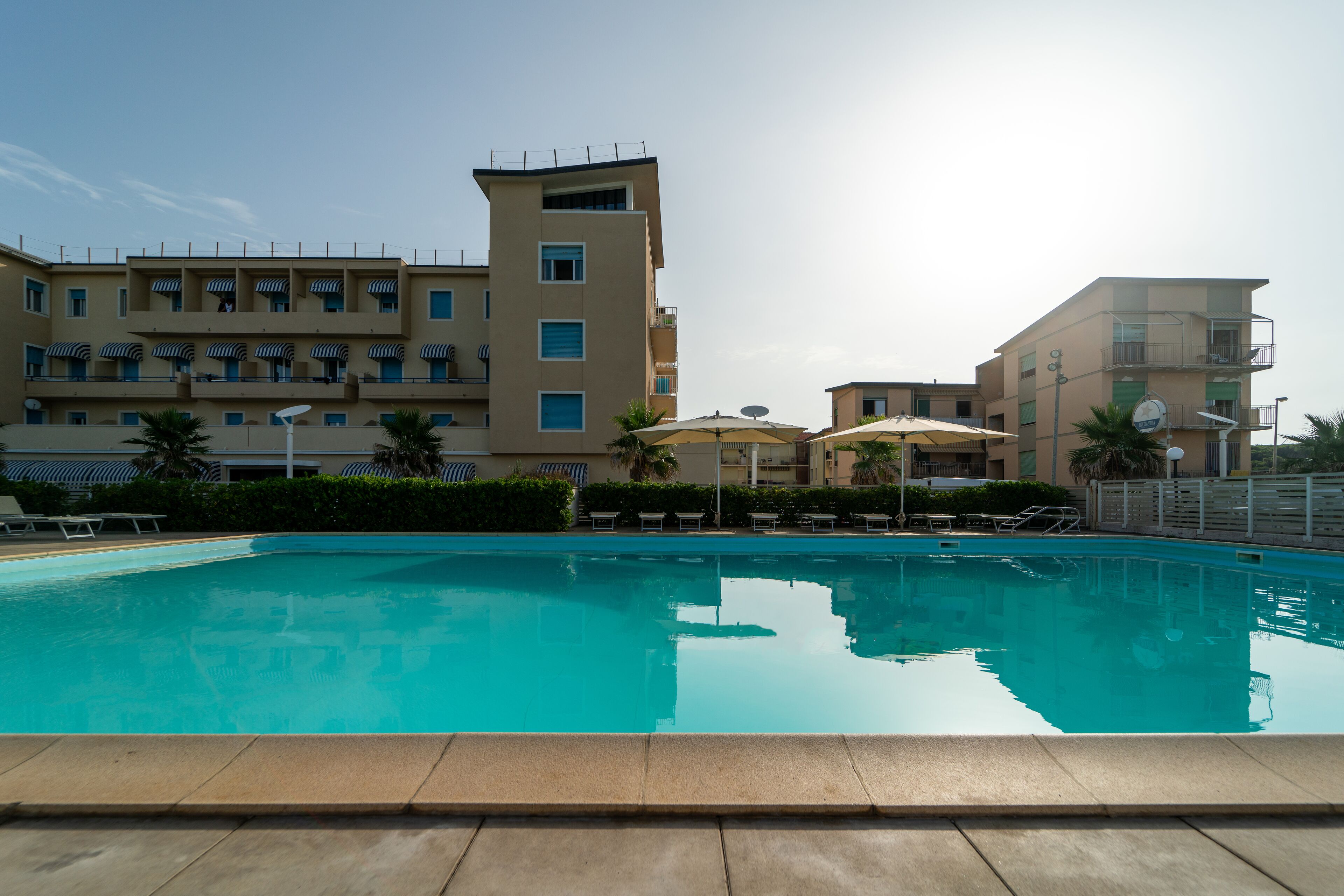 A swimming pool with lounge chairs and umbrellas, surrounded by multi-story buildings with balconies and satellite dishes.