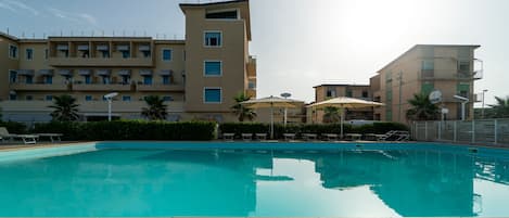 A swimming pool with lounge chairs and umbrellas, surrounded by multi-story buildings with balconies and satellite dishes.