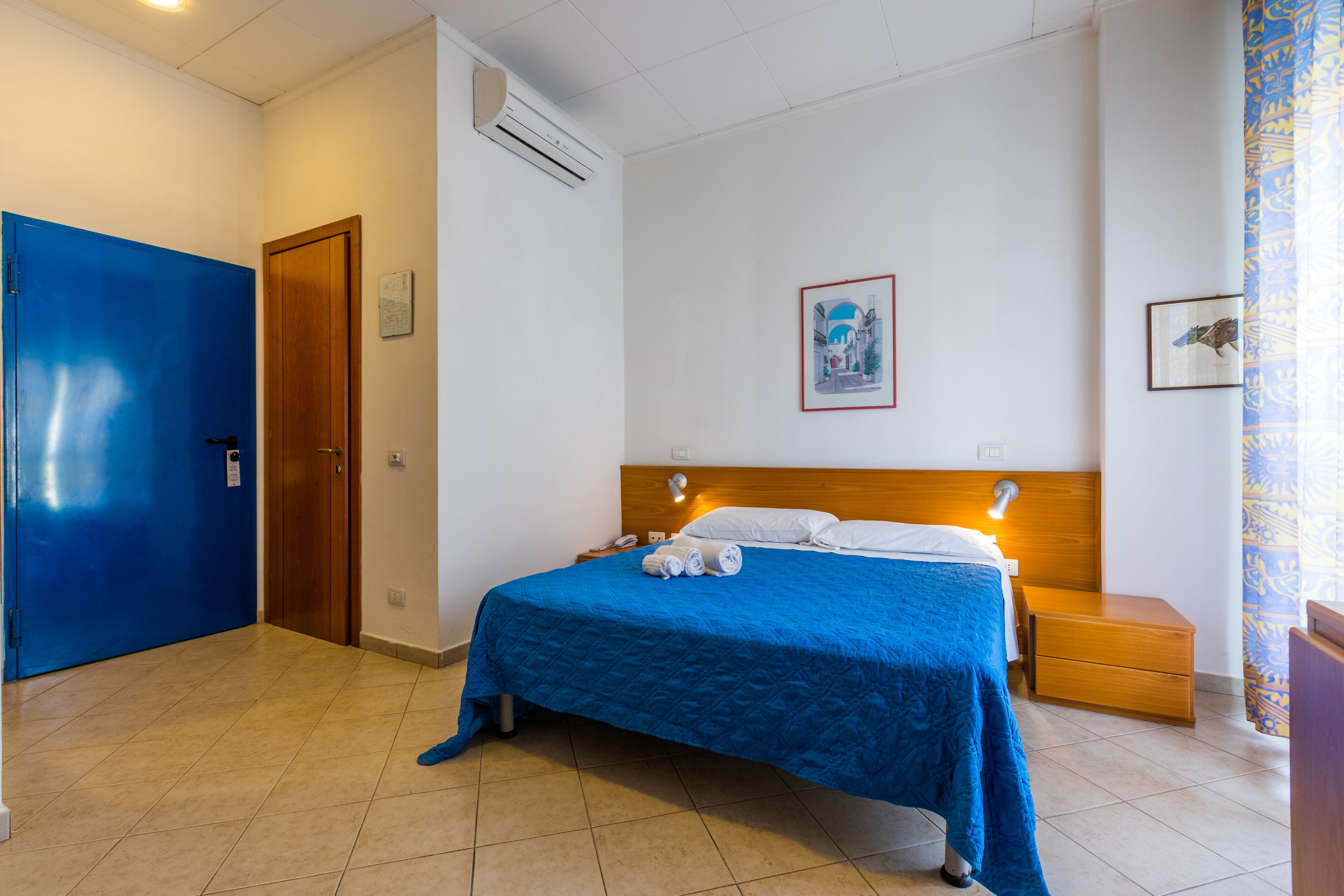 Ein Hotelzimmer mit einem großen Bett, einem Holzkopfende, einem Nachttisch, einer blauen Tür und einer wandmontierten Klimaanlage.